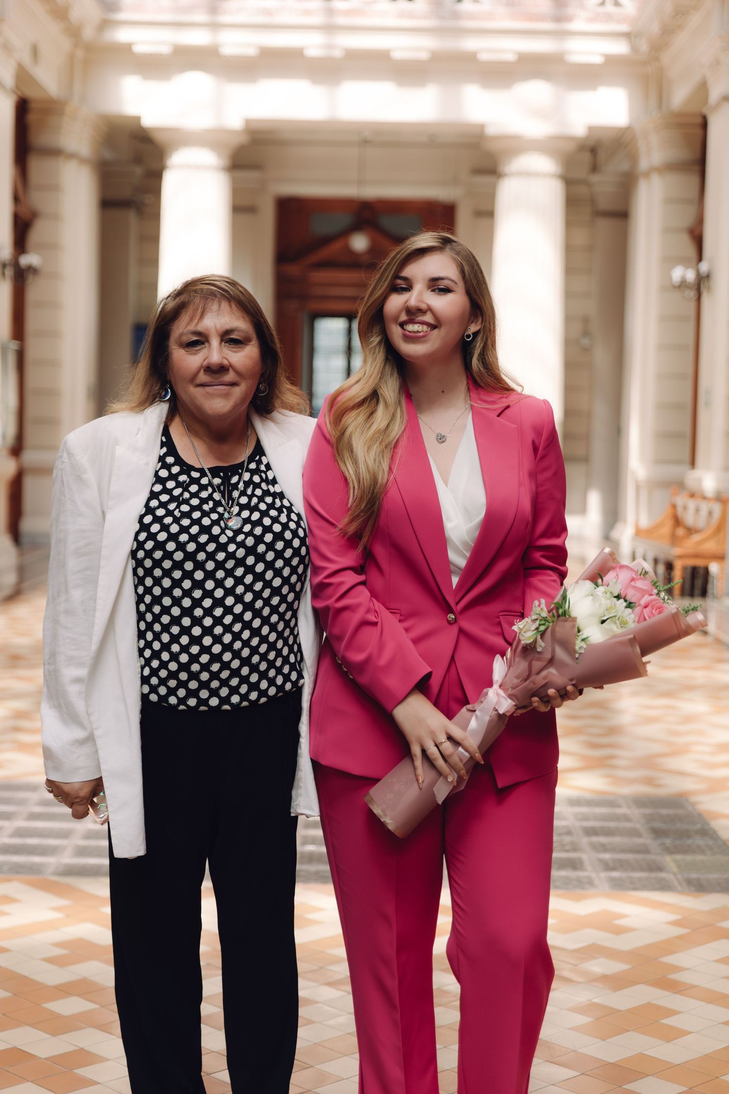 Two women smile together in grand hallway, one in pink suit holding a beautiful flower bouquet.