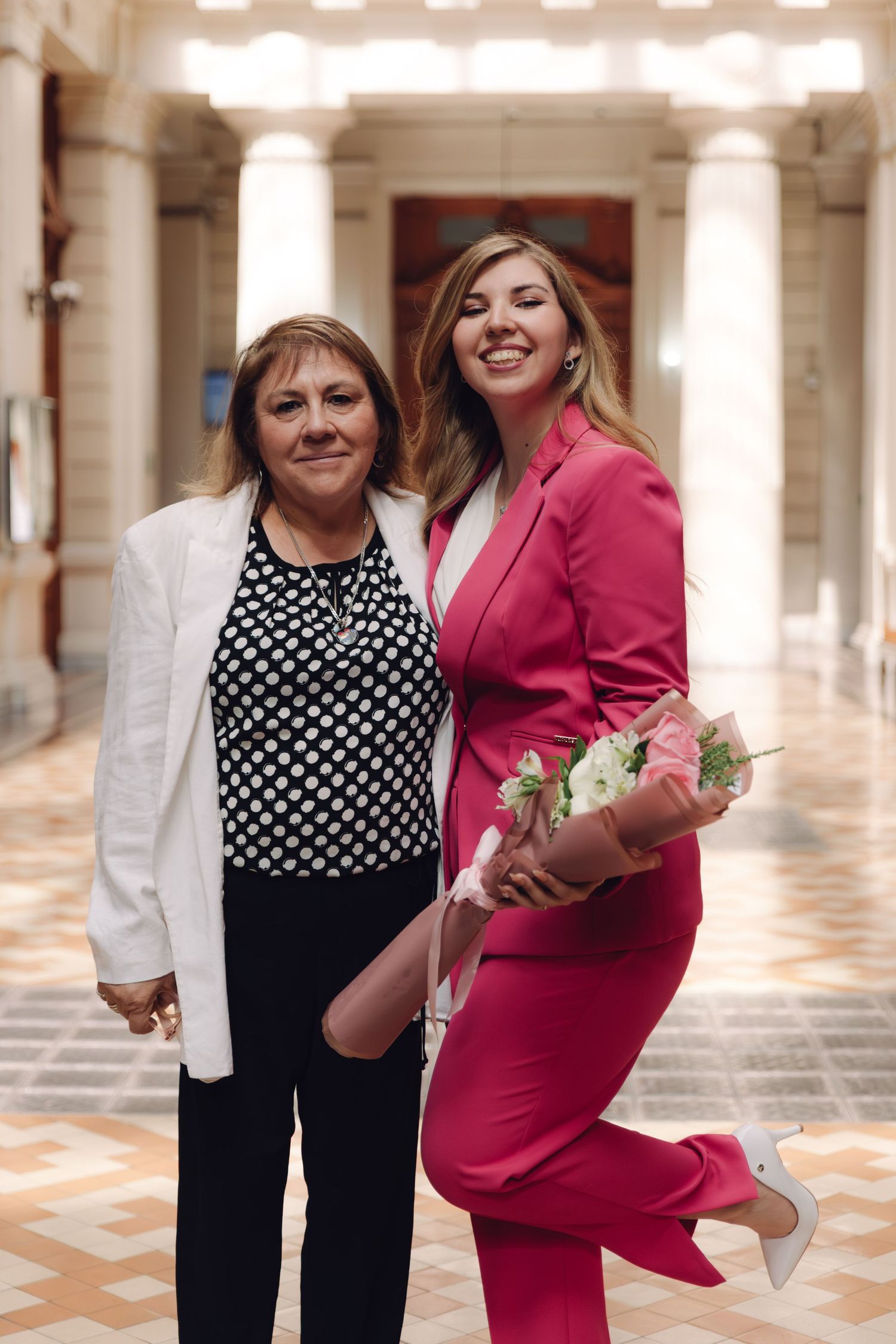 Two smiling women pose in ornate corridor, woman in pink pantsuit holds a pink floral bouquet.