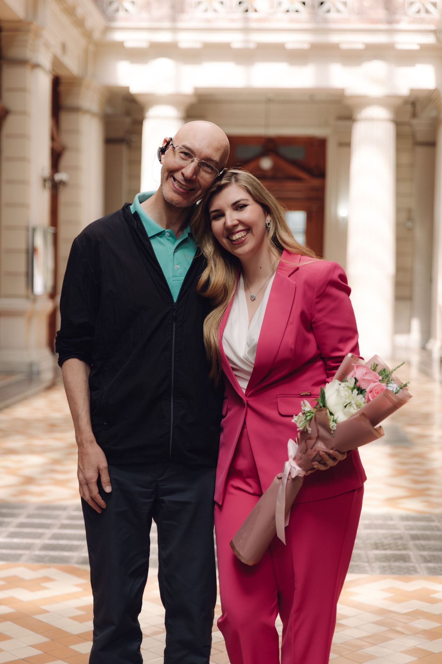 Bald man in casual wear and woman in pink suit smile together holding flowers in elegant hall.