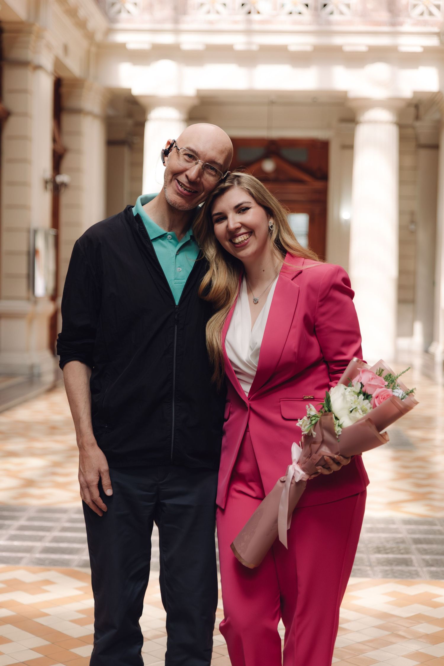 Smiling man and woman in pink suit with bouquet pose together in a grand columned hallway.