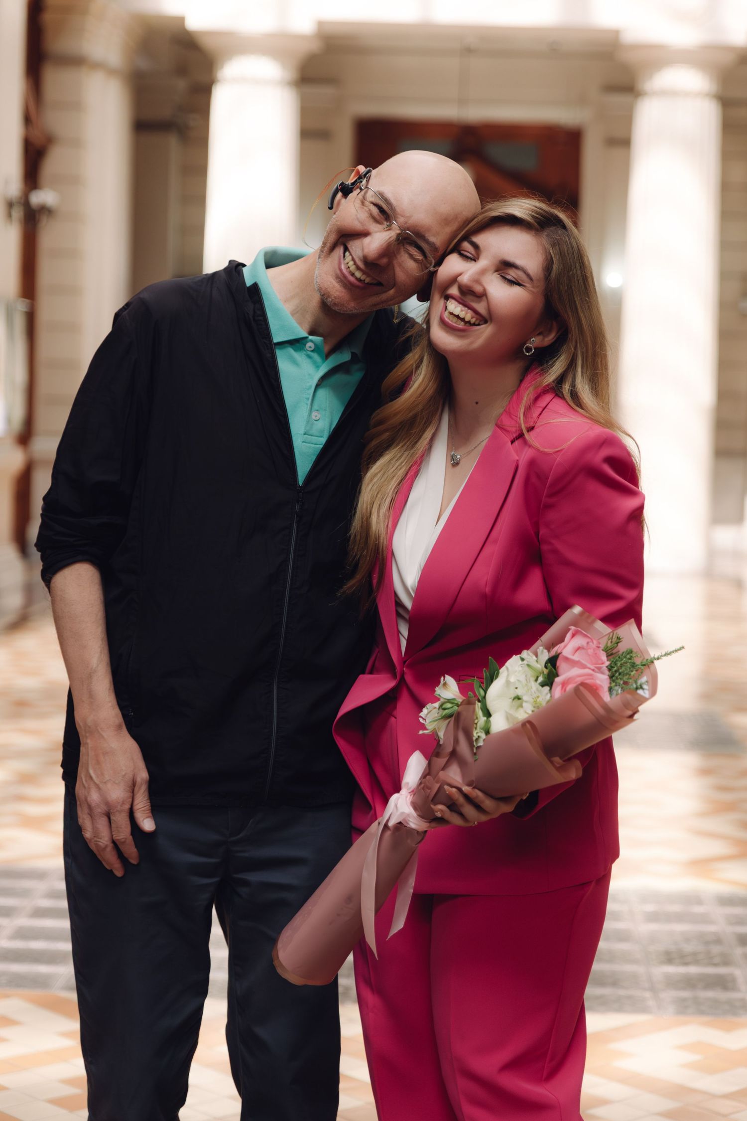 Smiling bald man and blonde woman in pink suit embracing while holding flower bouquet in elegant hallway.
