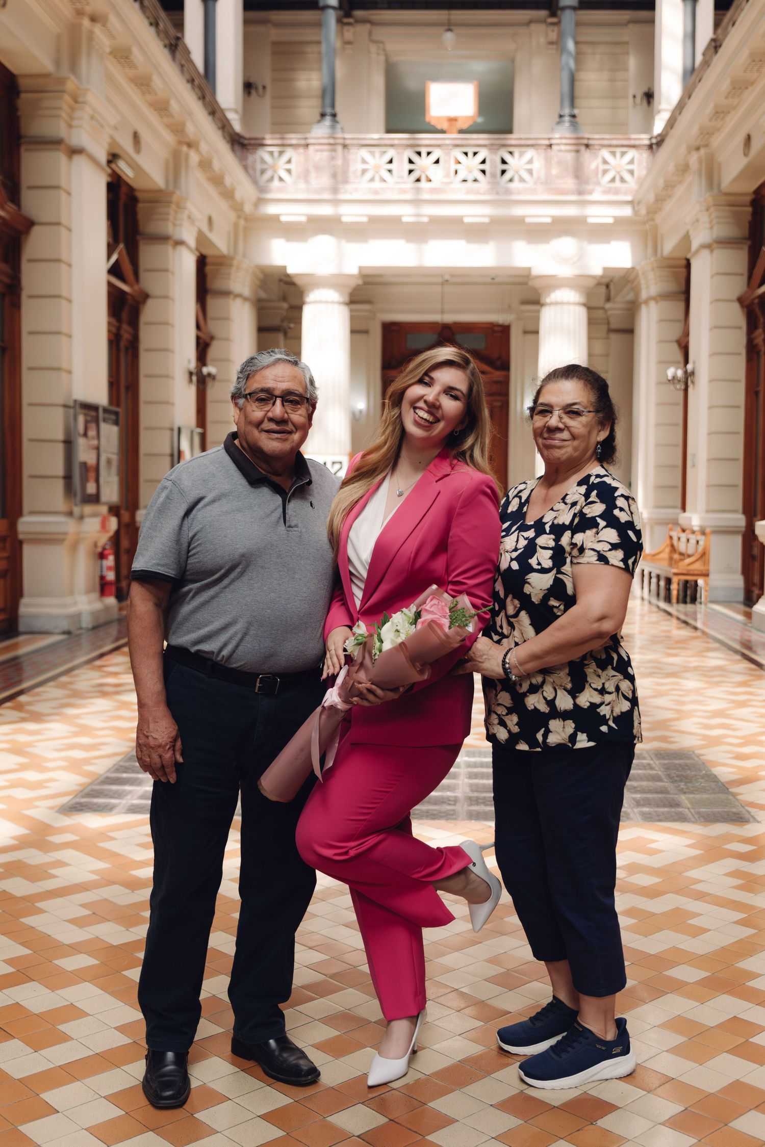 Blonde woman in pink suit holding bouquet poses with older couple in ornate building corridor.