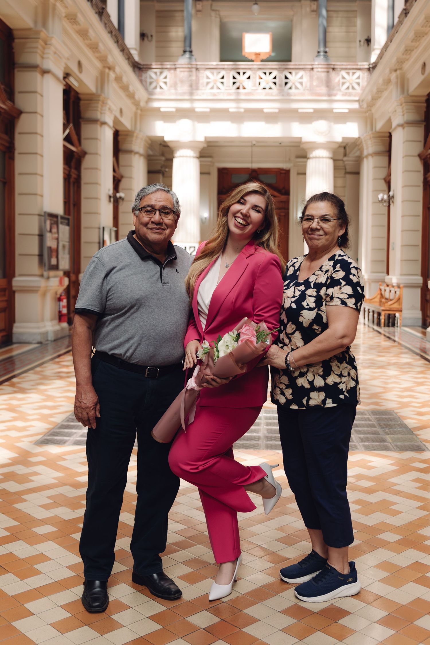 Young woman in pink pantsuit smiles with elderly couple in decorative indoor hallway with columns.