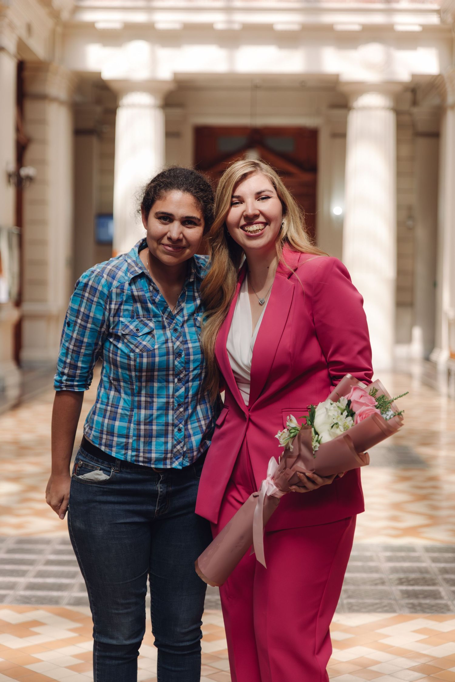 Two smiling women pose together in grand columned hall, one in pink suit holding flower bouquet.