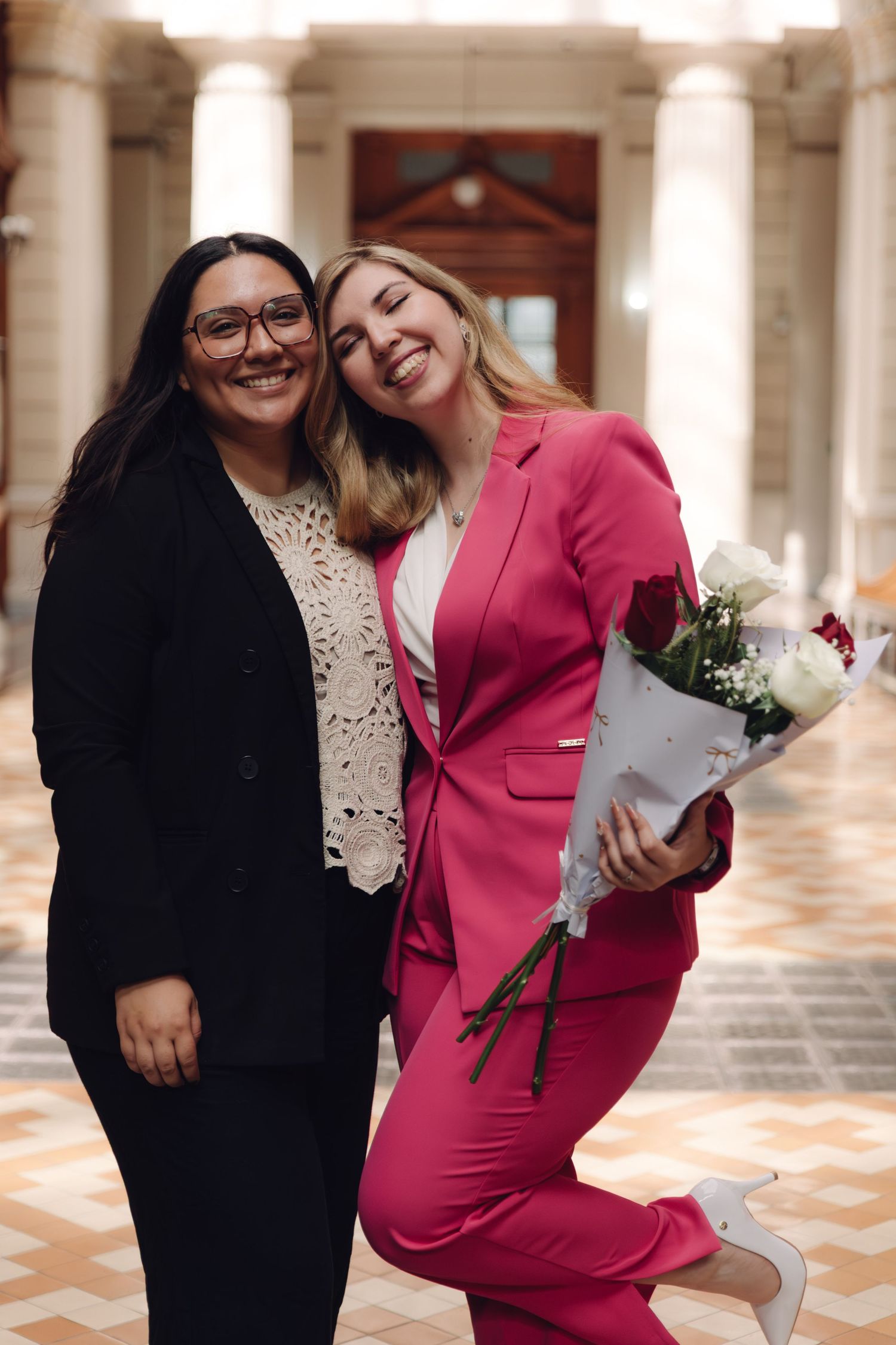 Two happy women posing together in columned hallway, one in pink suit holding bouquet of roses.