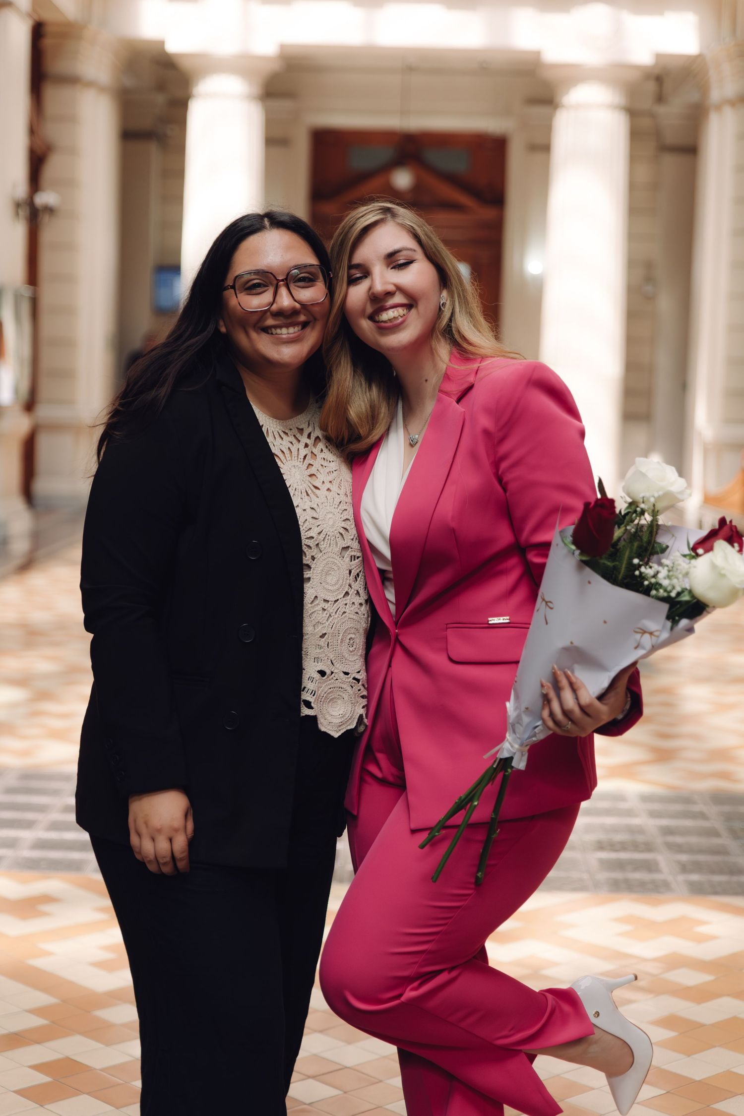 Two women smiling and embracing in elegant corridor, one in pink suit holding red and white roses.