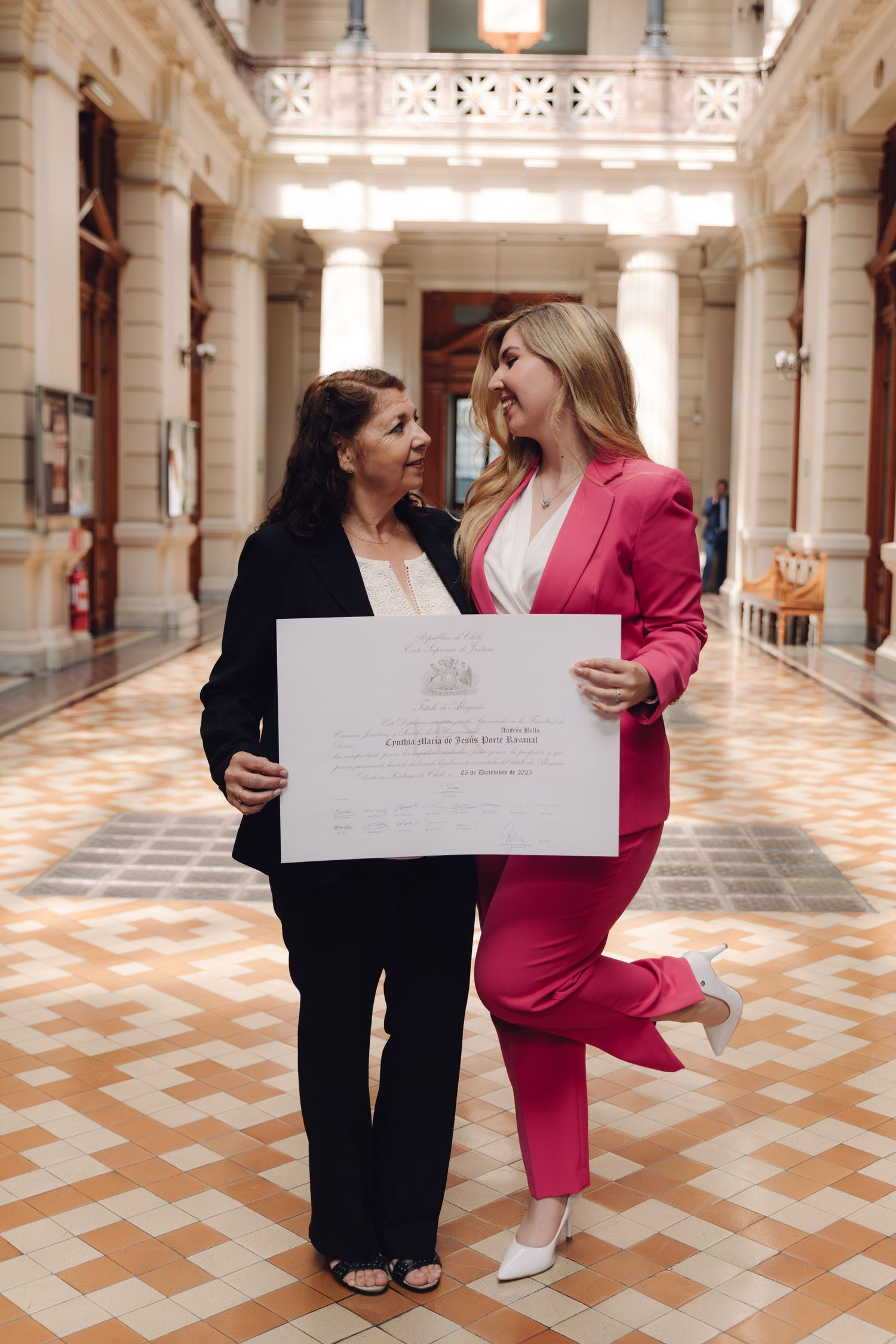 Two women in elegant hall proudly displaying official diploma certificate while smiling at each other.