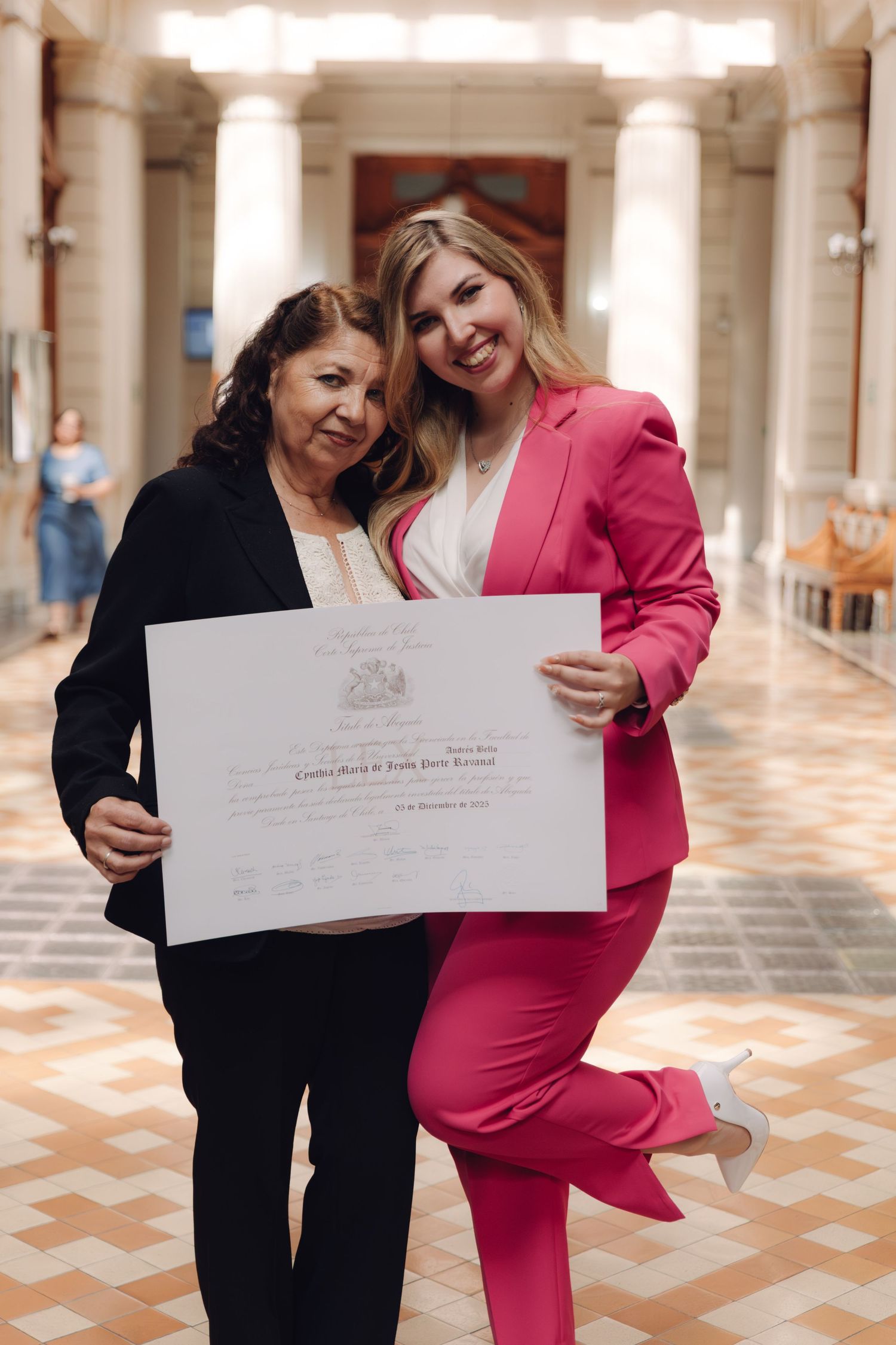 Smiling woman in pink suit and older woman proudly hold official diploma certificate in grand hall.