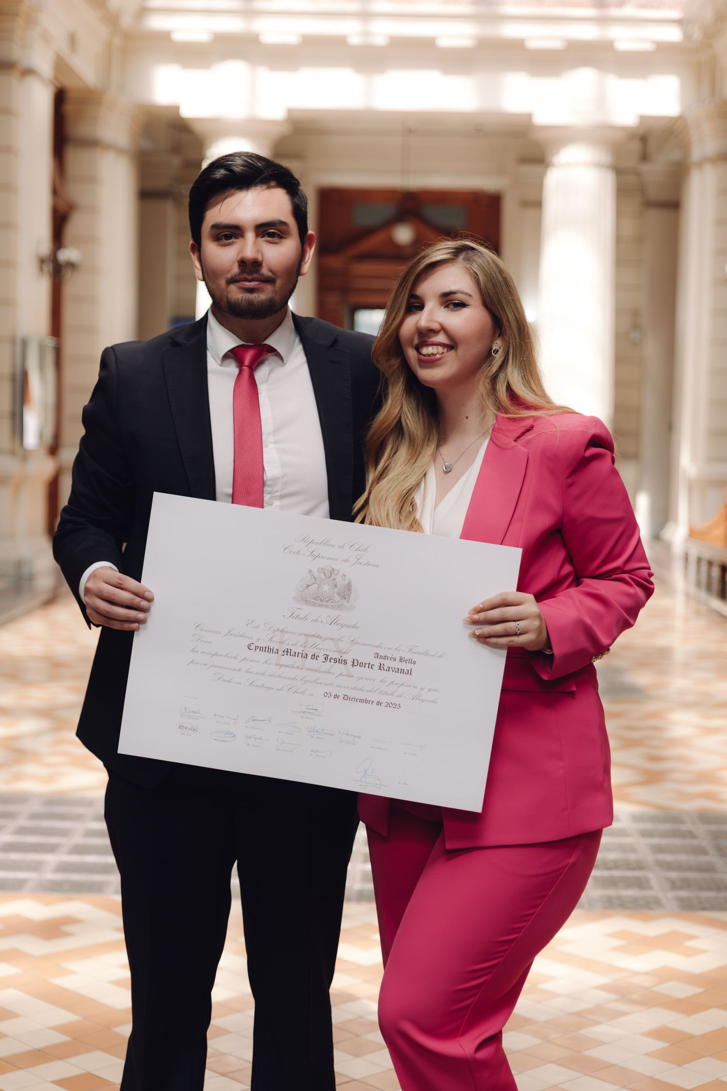 Young man in black suit and woman in pink pantsuit proudly display official diploma in elegant hall.