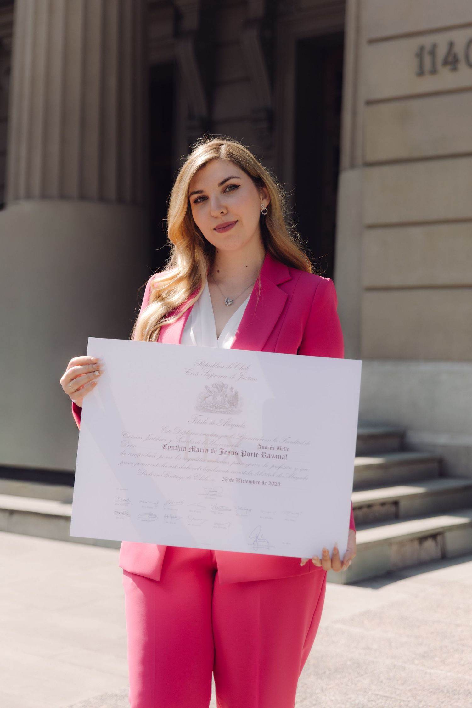 Proud young blonde woman in pink suit holding official diploma certificate outside building entrance.