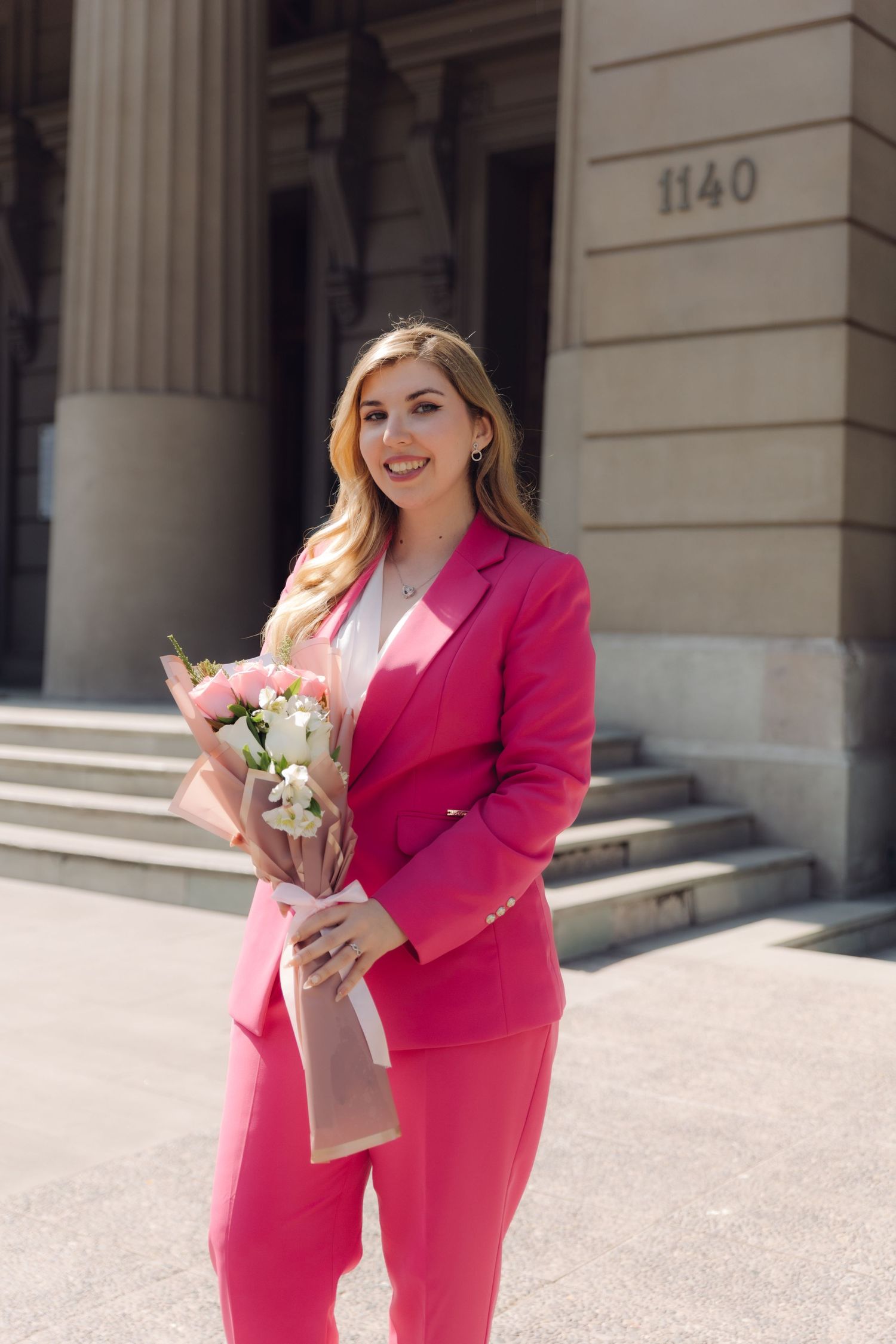 Woman in pink suit smiling and holding a bouquet of flowers outside a building.