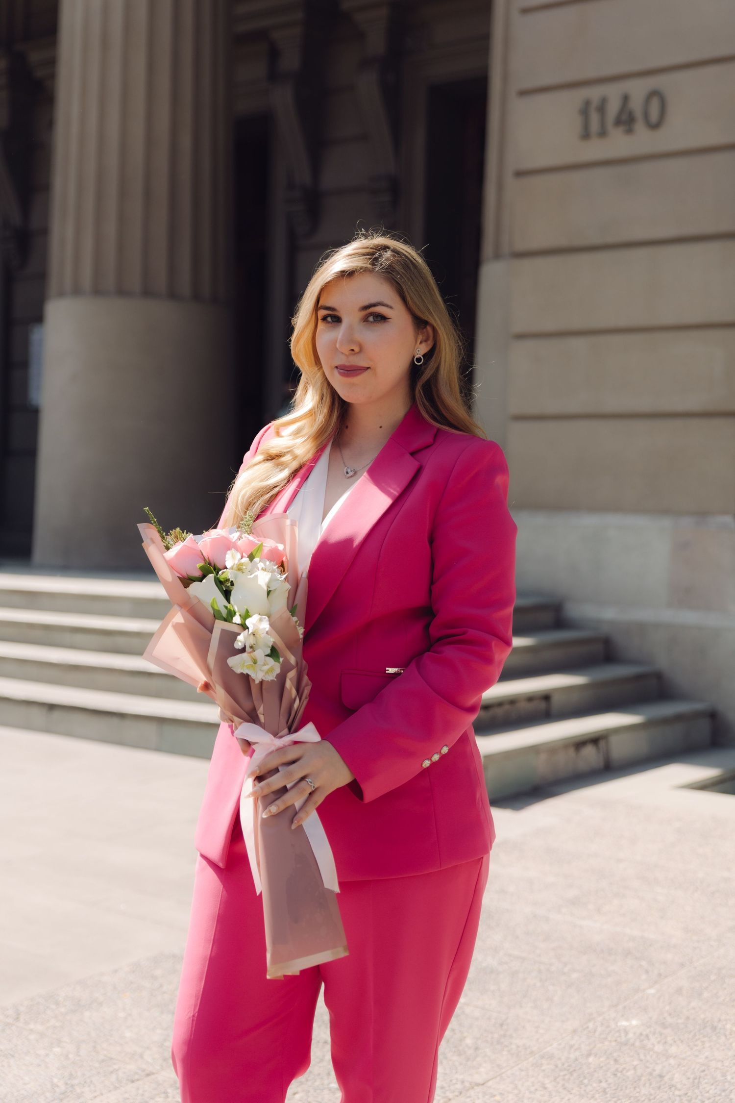 Blonde woman in hot pink blazer and pants holding a floral bouquet outdoors.