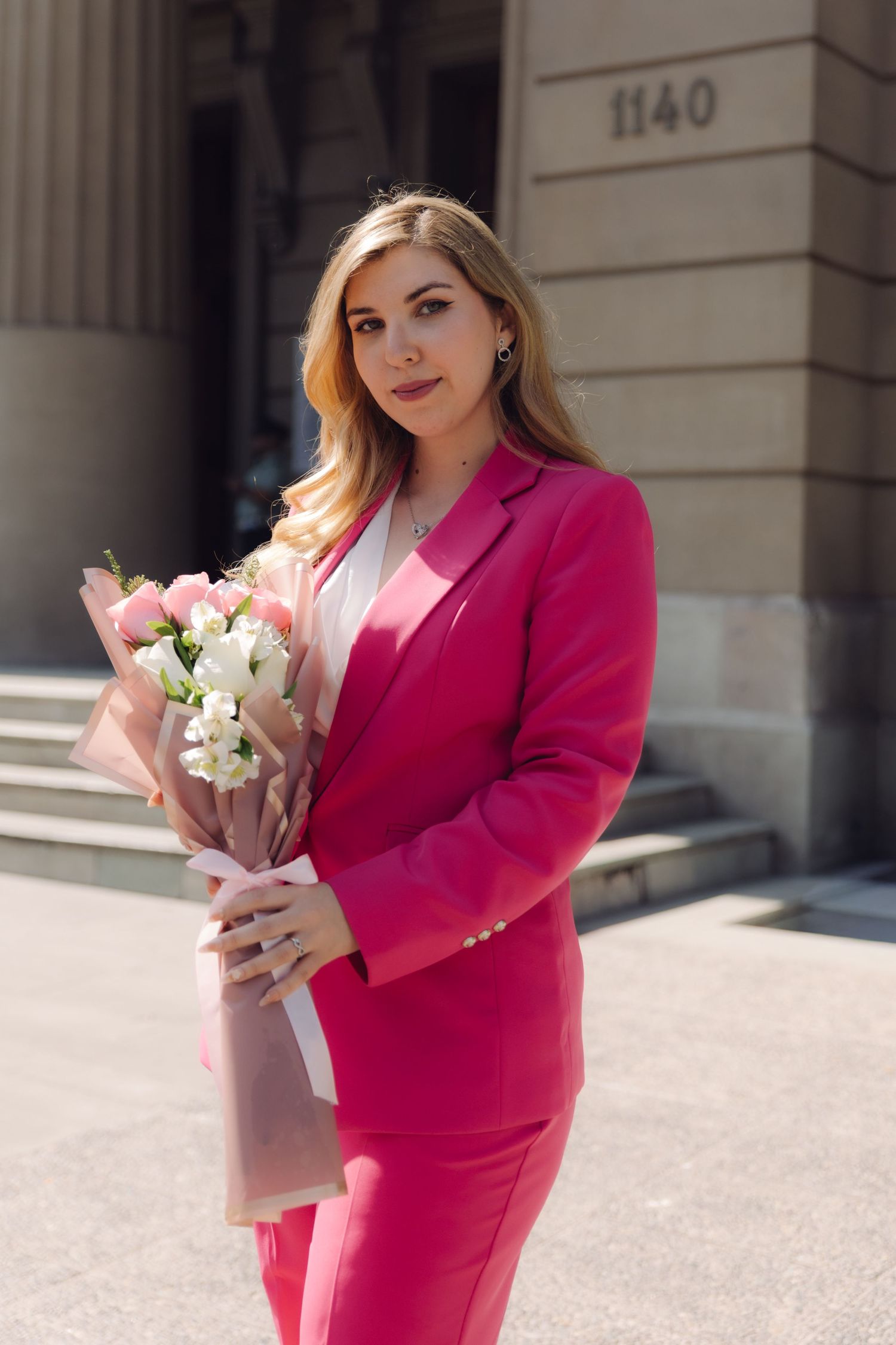 Elegant woman in pink suit holding white and pink flower bouquet near building.