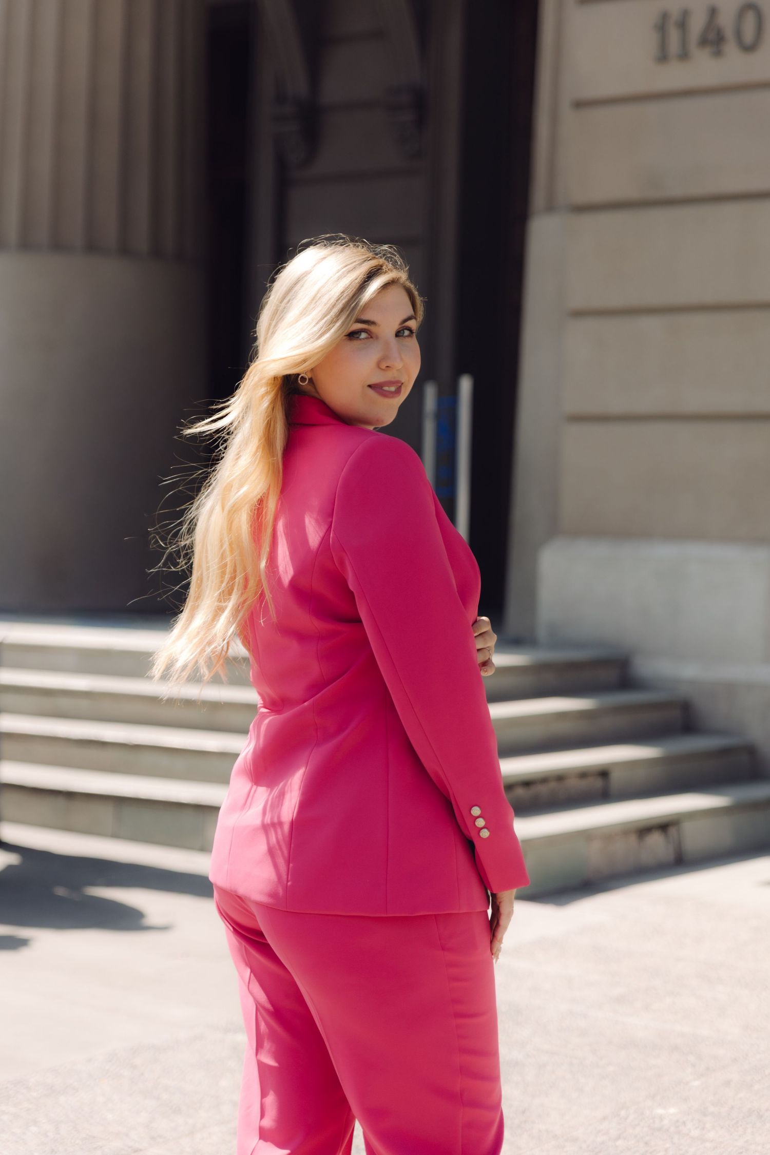 Blonde woman in bright pink suit walking away from stone building steps.
