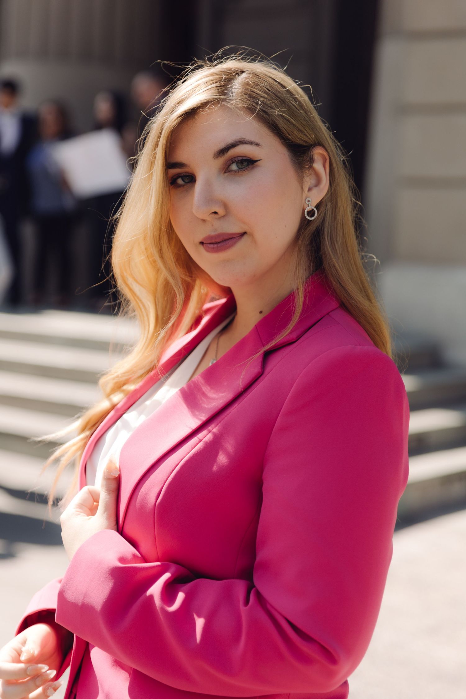 Young woman in pink blazer with arms crossed posing outside stone building.