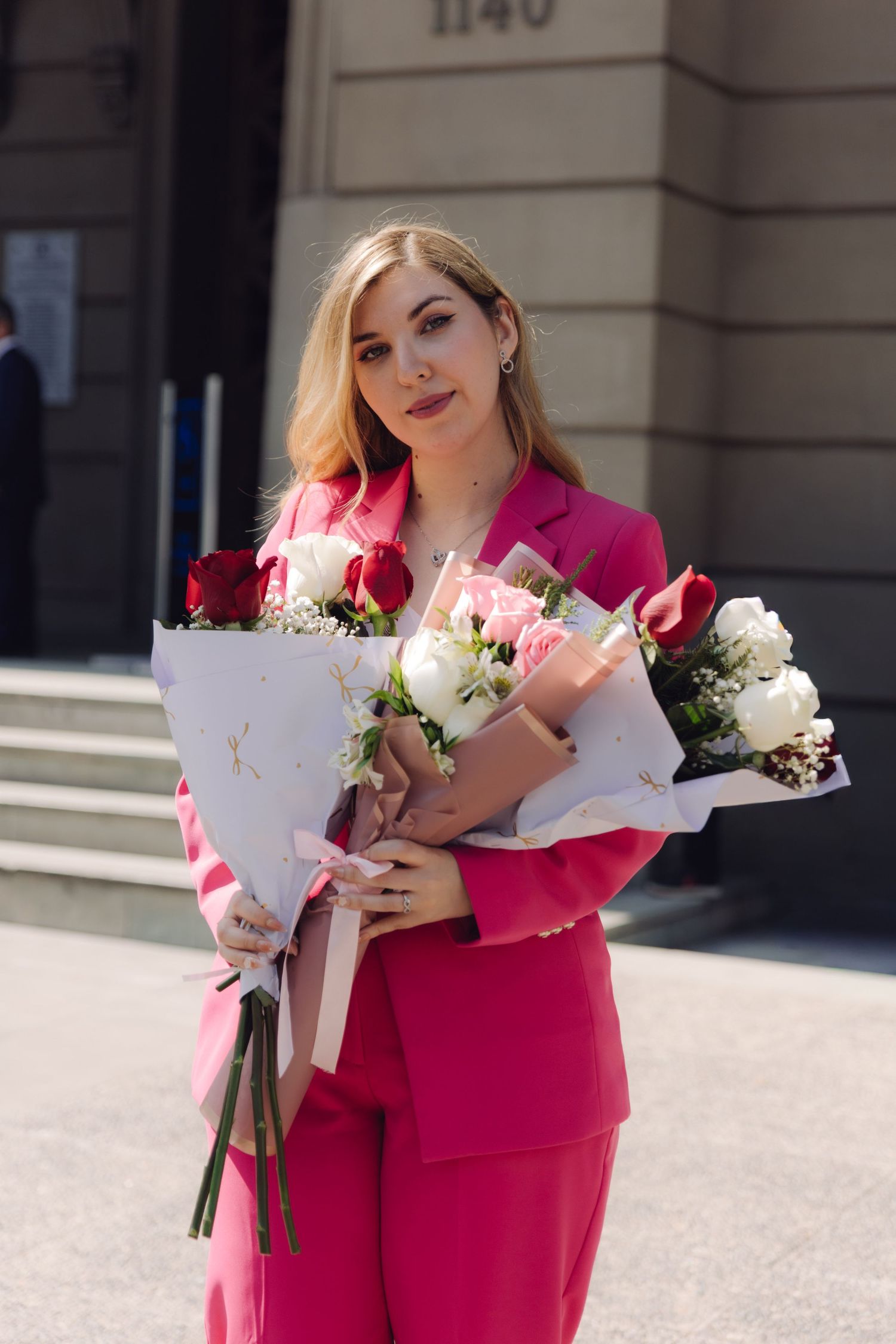 Smiling woman in pink suit holding two large flower bouquets outside building.