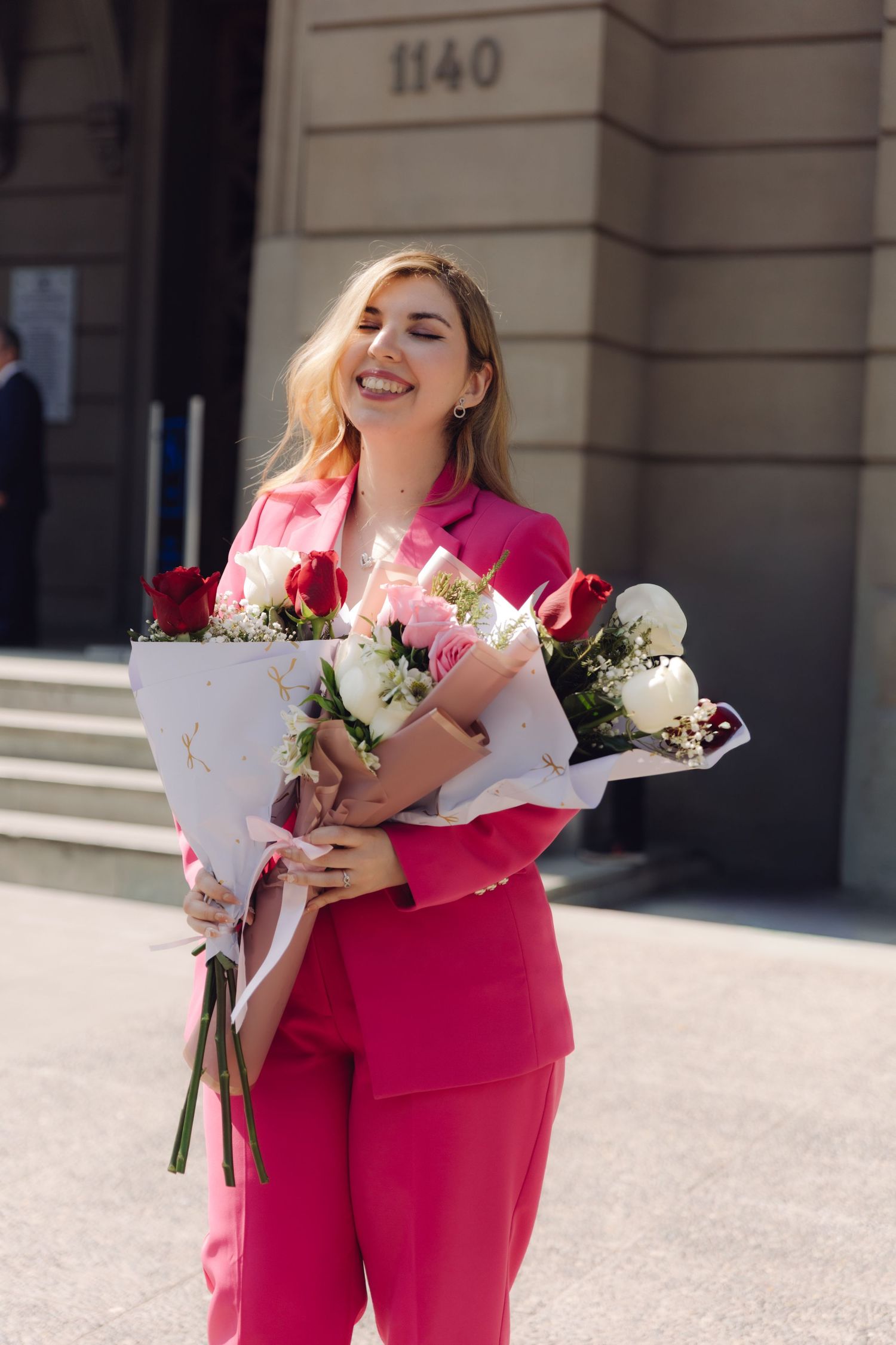 Happy woman in pink pantsuit holding two bouquets of roses outdoors.