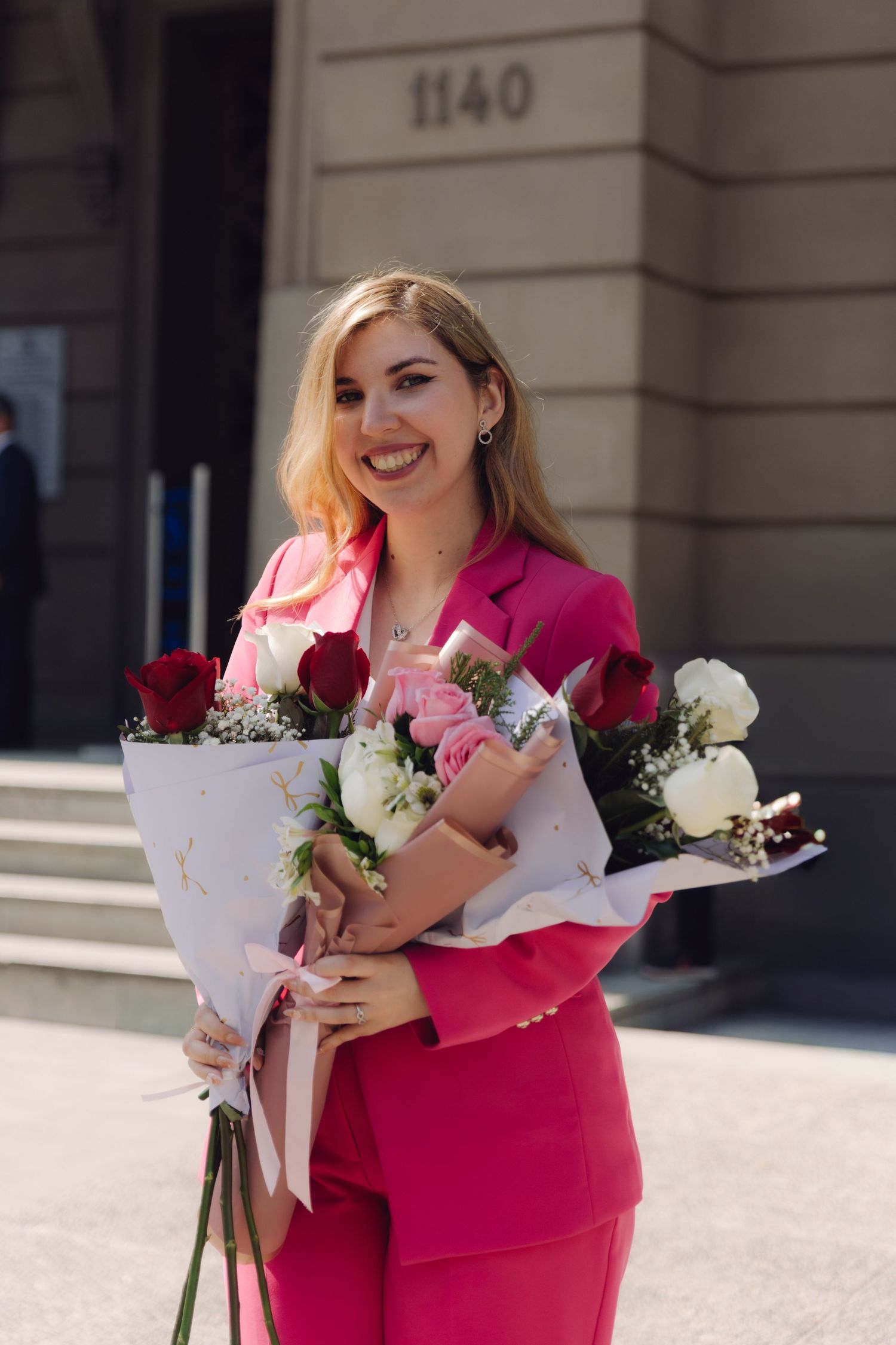Cheerful blonde woman in pink suit holding two floral bouquets outside.