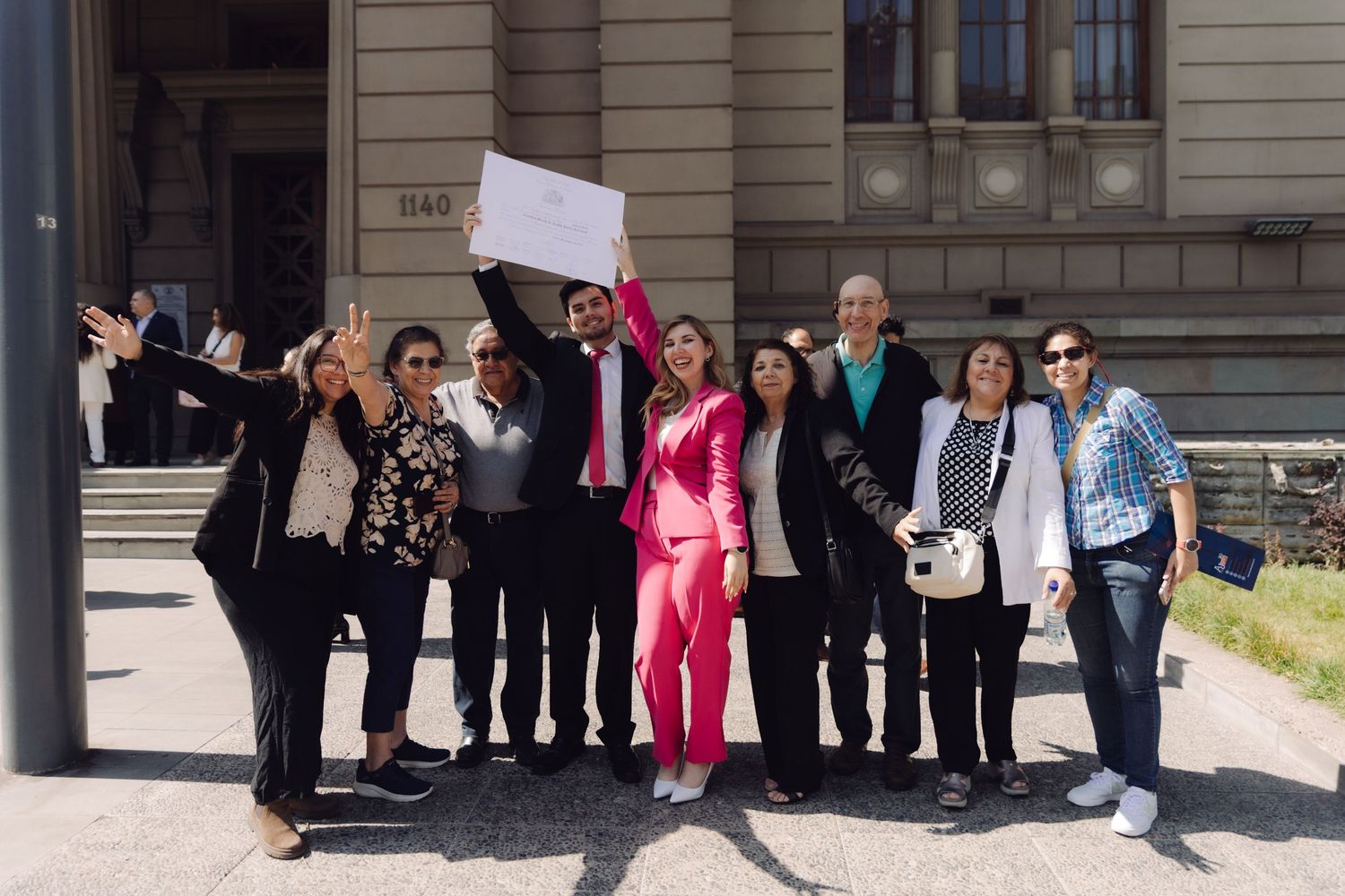 Celebrating group of people posing outside a building with a raised document.