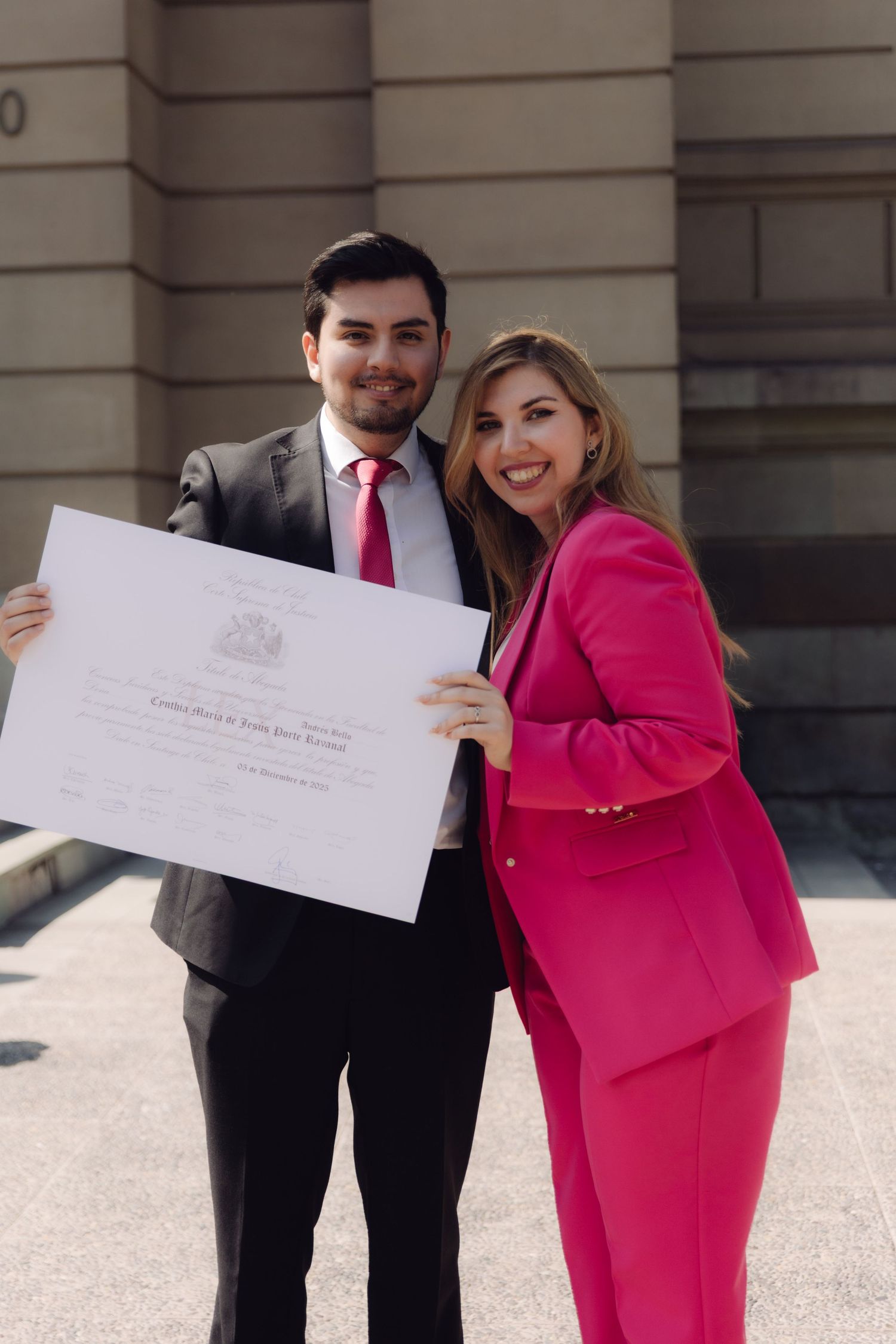 Man in suit and woman in pink blazer proudly holding a diploma certificate outdoors.