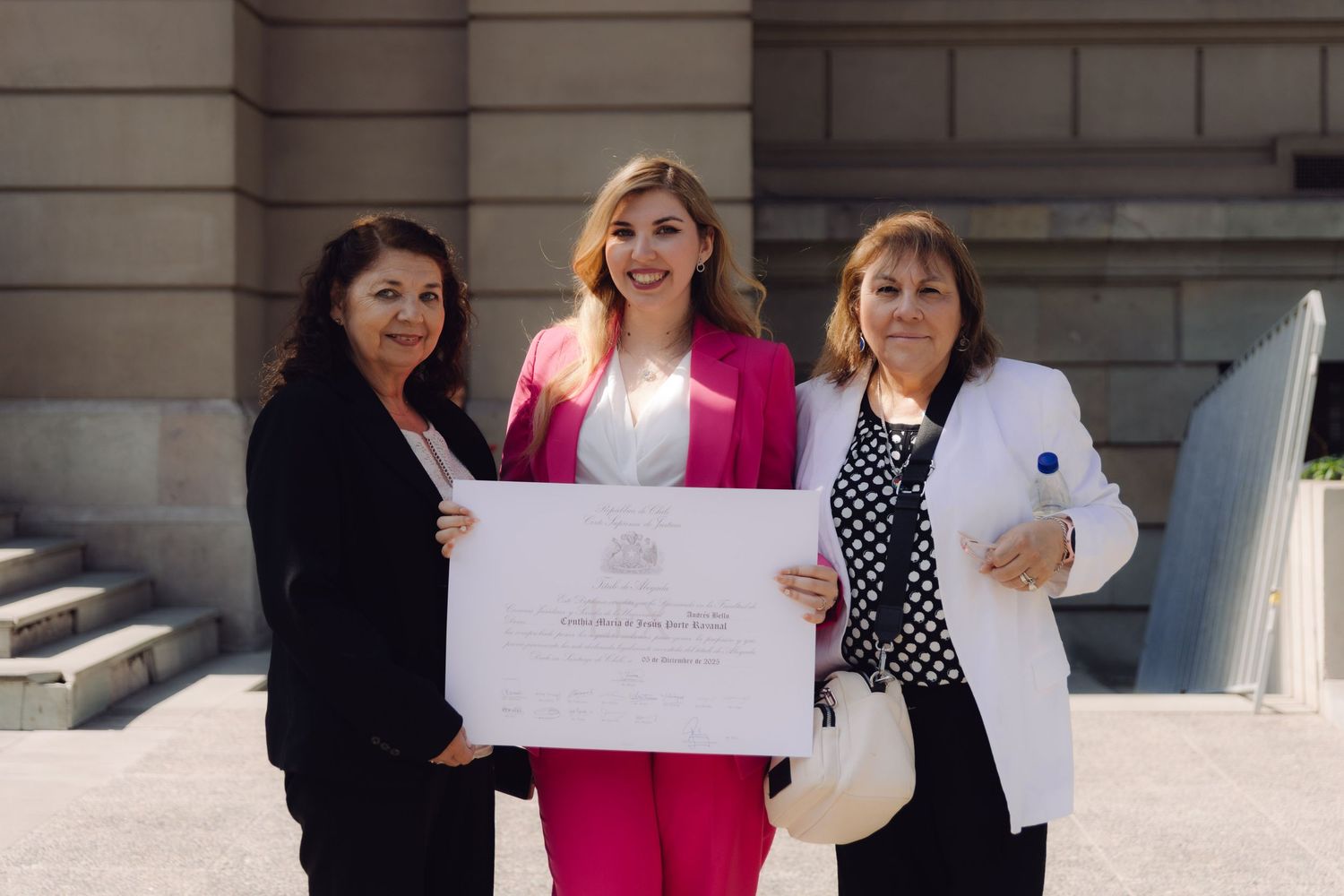 Three women in formal attire holding a diploma certificate outdoors at a ceremony.