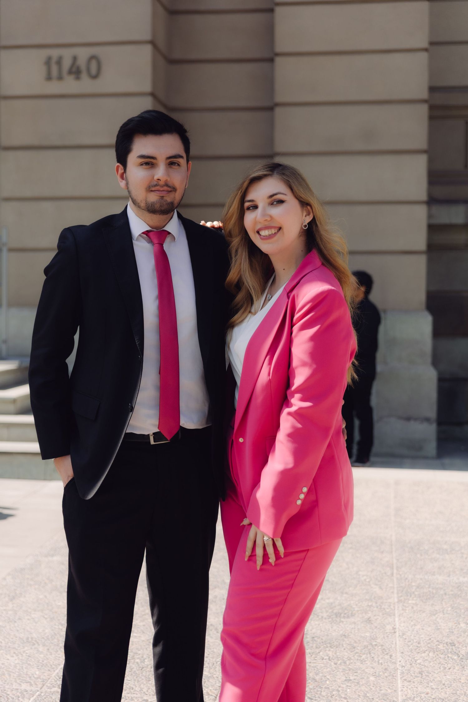 Young man in black suit and woman in pink blazer posing together outside building 1140.