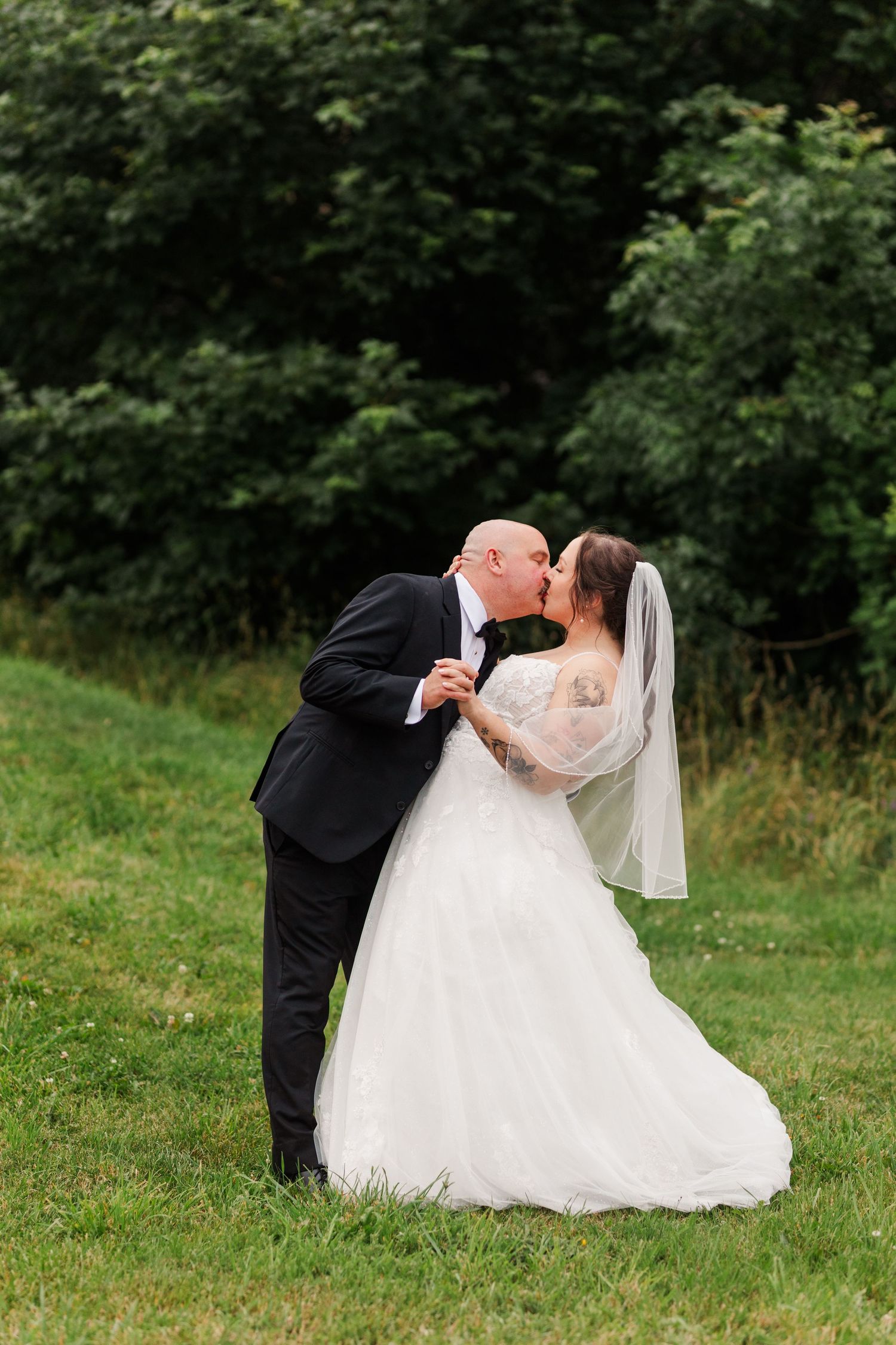 A bride and groom share a romantic kiss on a green lawn with lush trees in the background on their wedding day.