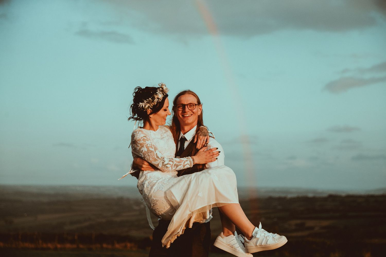 Couple celebrating wedding with rainbow in background, one partner lifting the other against scenic landscape.