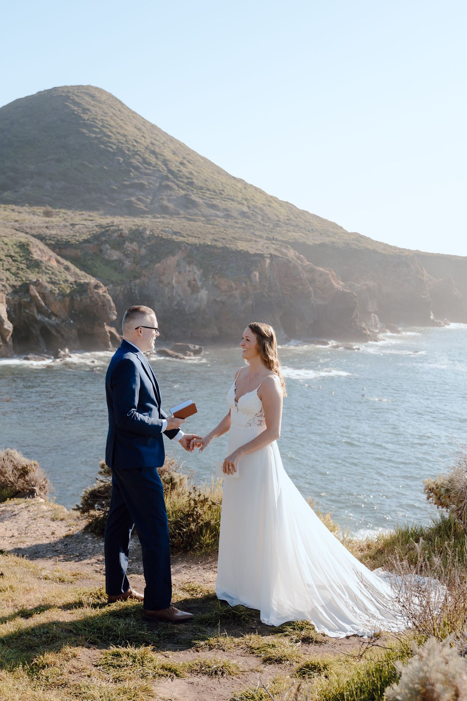 Couple exchanging vows at coastal cliff wedding with ocean and mountain views on a sunny day.