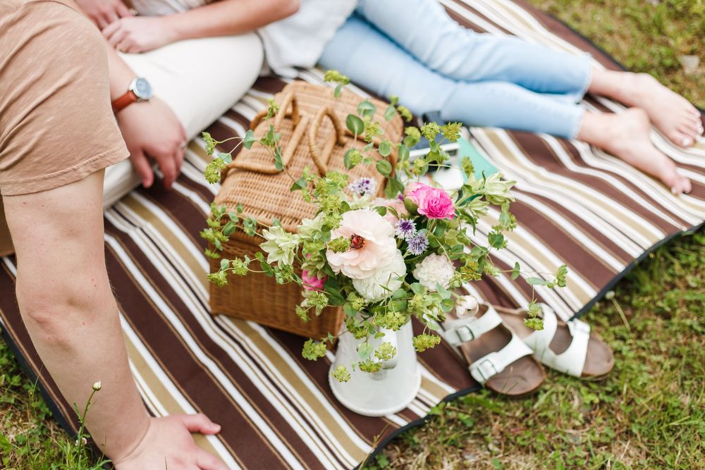 Picnic blanket with floral bouquet, wicker basket, and sandals on grass with people sitting nearby.
