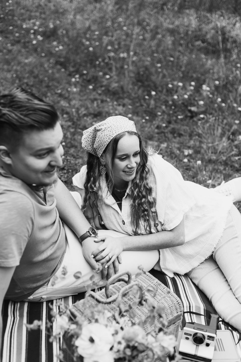 Black and white photo of a young couple enjoying a romantic picnic outdoors on a blanket with a basket and camera.