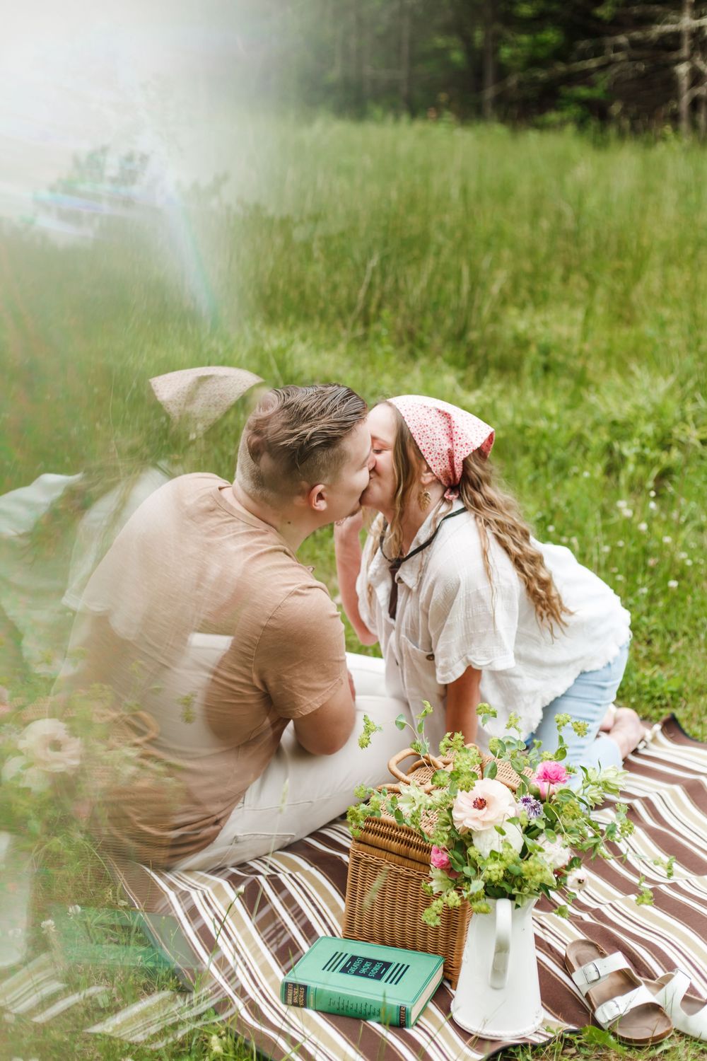 Couple sharing a romantic kiss during a countryside picnic on a striped blanket with flowers and a wicker basket.