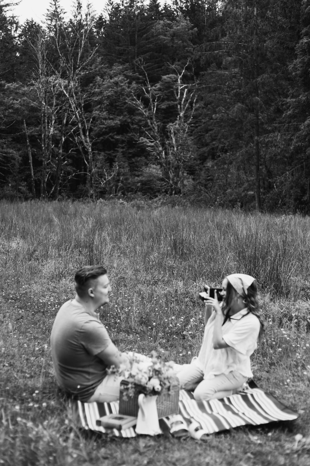 Black and white photo of a couple enjoying a romantic picnic in a meadow, woman photographing man with vintage camera.