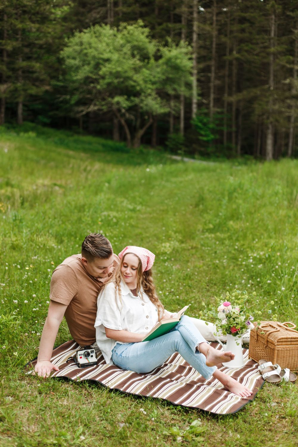 Couple sitting on a picnic blanket in a green meadow, woman in pink hat reading a book while leaning on her partner.