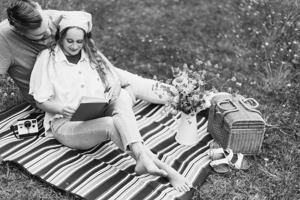 Black and white photo of a couple enjoying a romantic picnic on a striped blanket with flowers and a wicker basket.