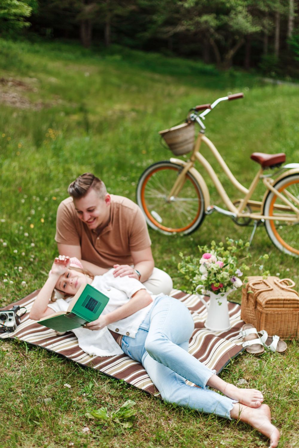 Couple relaxing on a picnic blanket outdoors, woman lying down reading a book while man sits beside her near a bicycle.