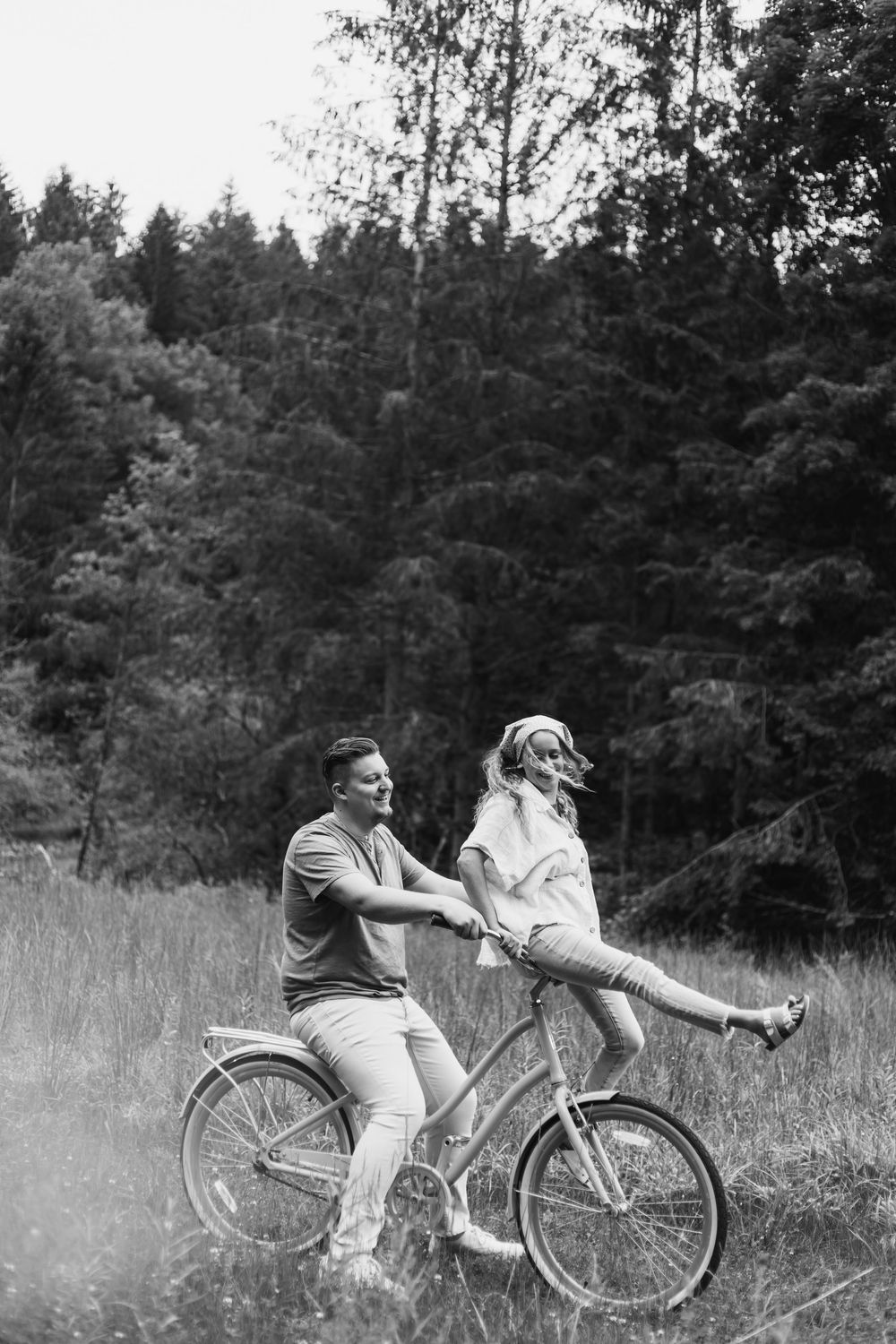 Black and white photo of a couple sharing a bicycle ride in a meadow with a forest backdrop.