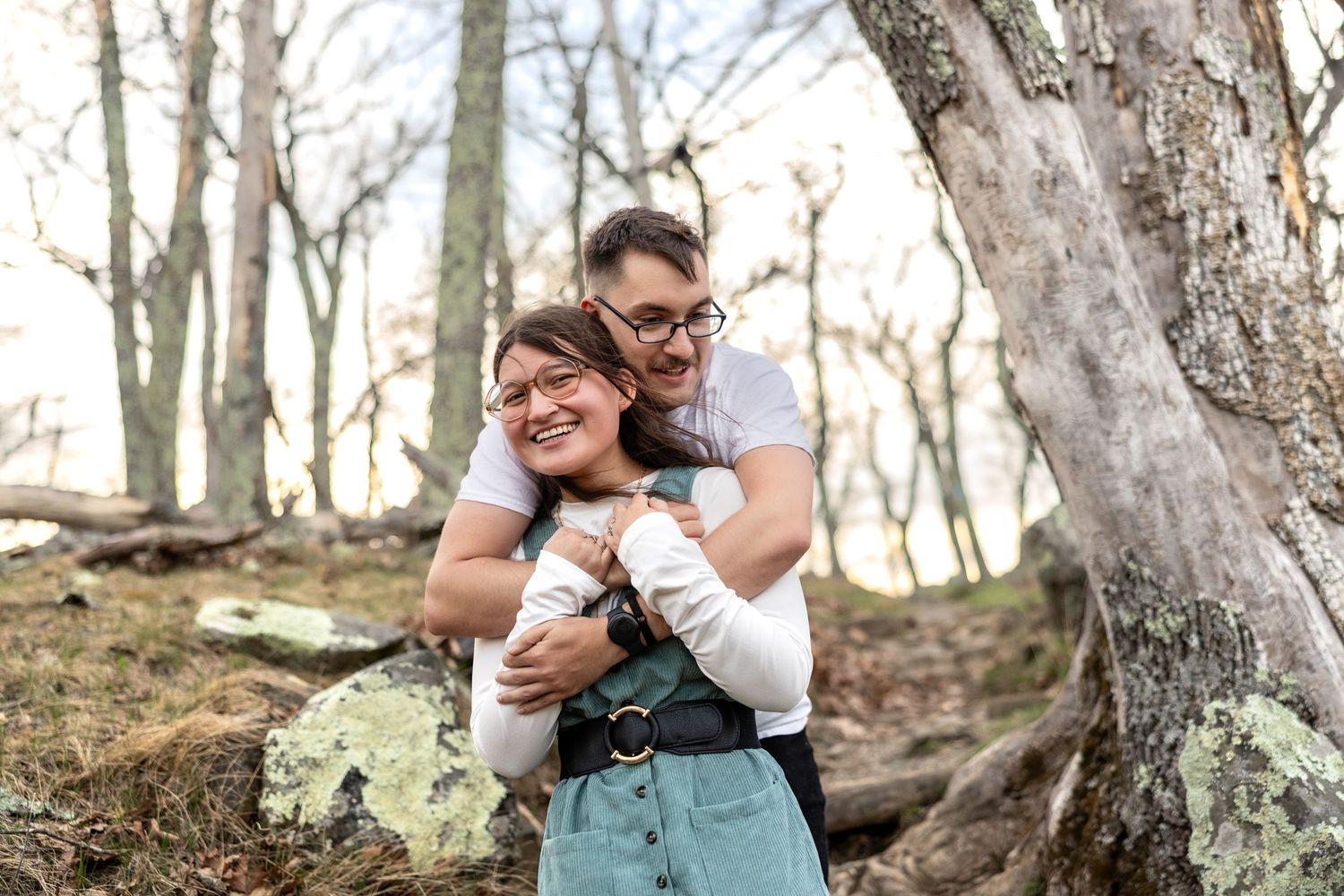 A joyful couple embraces while standing in a forest setting with bare trees and rocky terrain in the background.