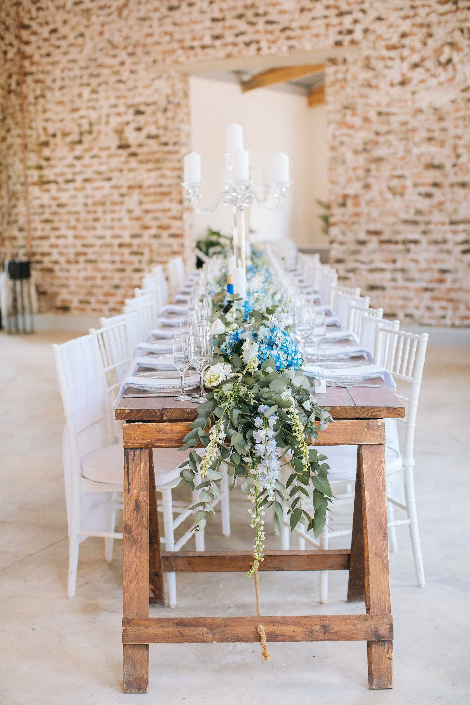 Rustic wooden banquet table with blue floral garland, white candelabra, and chiavari chairs in a brick venue.