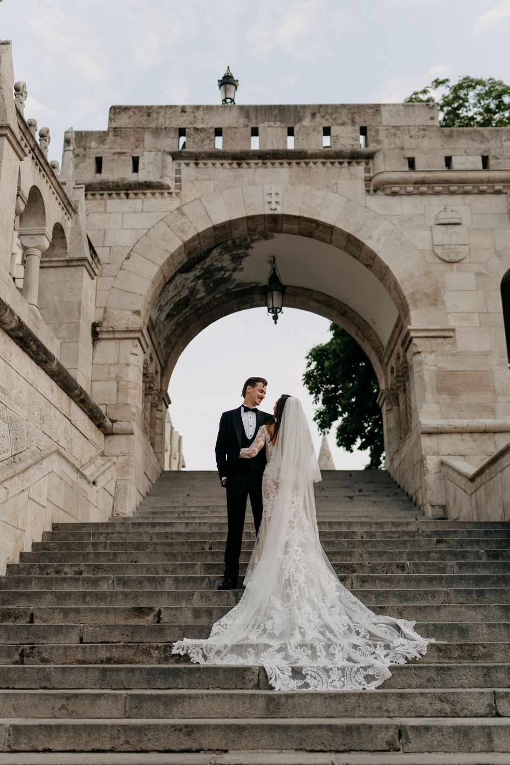 Bride and groom standing under a stone arch on castle steps with flowing lace cathedral veil.