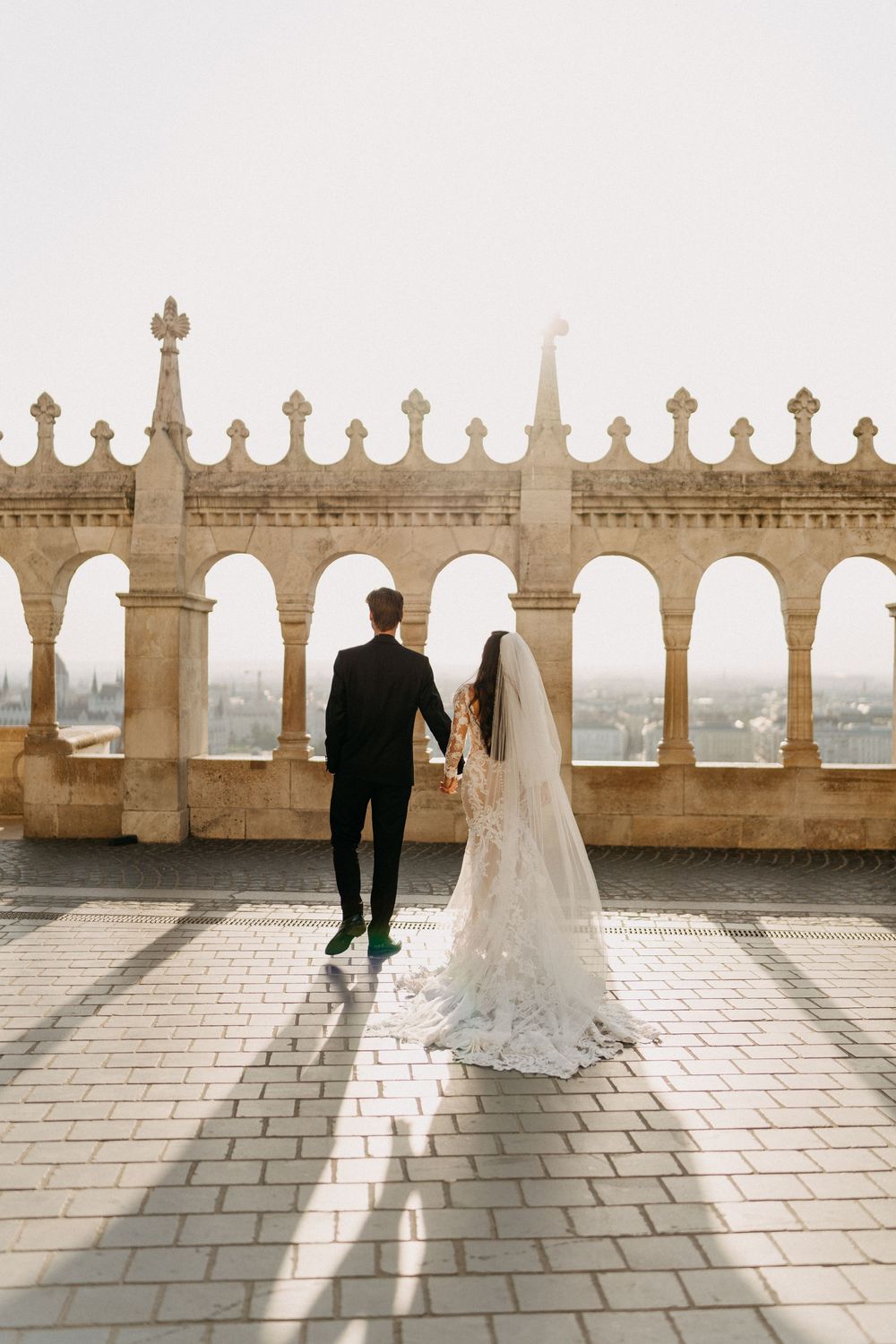 Bride and groom holding hands walking toward ornate stone balustrade overlooking Budapest cityscape.