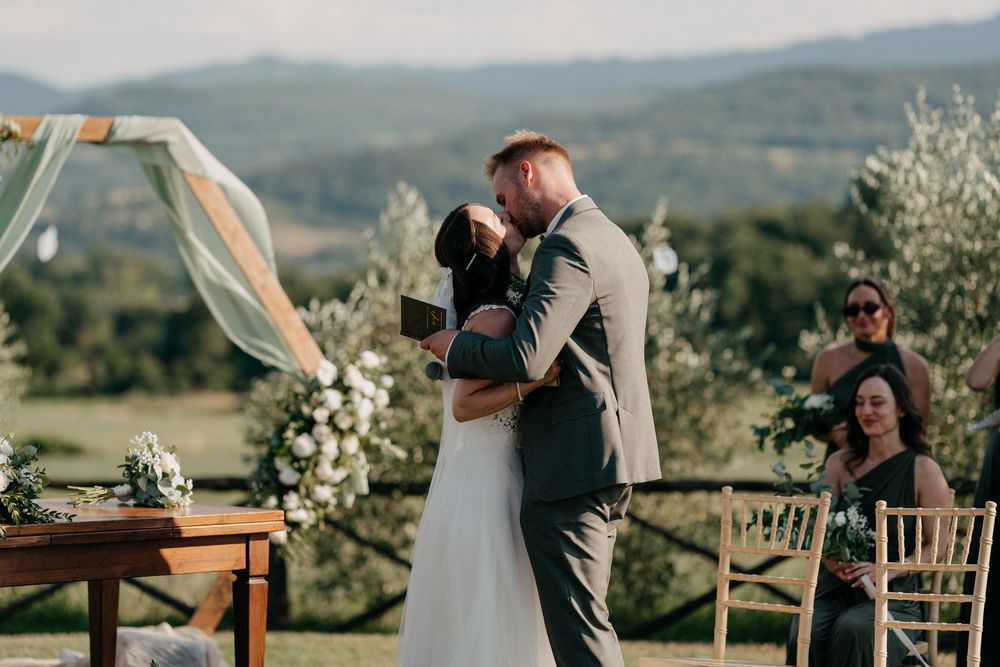 Bride and groom share their first kiss at an outdoor wedding ceremony with rolling Tuscan hills behind them.