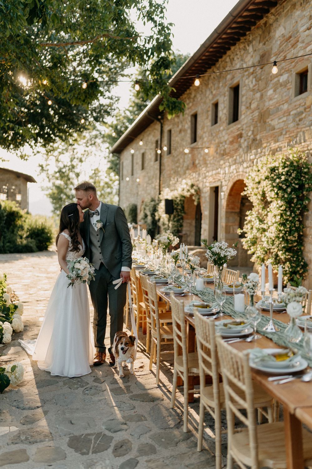Bride and groom share a kiss beside an elegantly set long reception table at a Tuscan stone farmhouse.