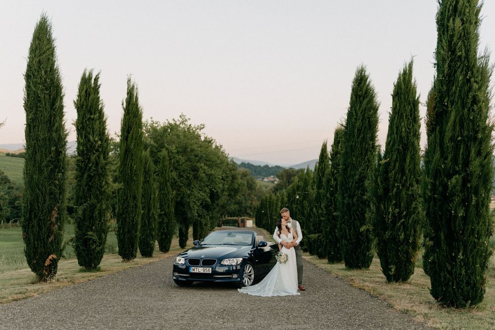 Bride and groom pose beside a black convertible car on a cypress-lined driveway at dusk in Tuscany.