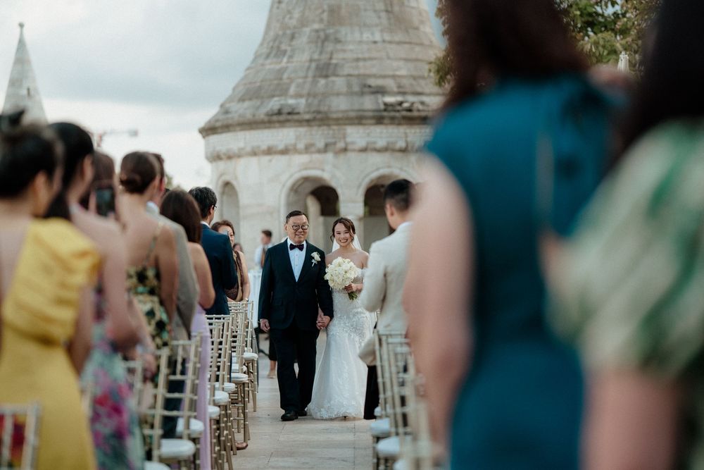 Bride walking down aisle with escort at outdoor castle wedding ceremony in Budapest.