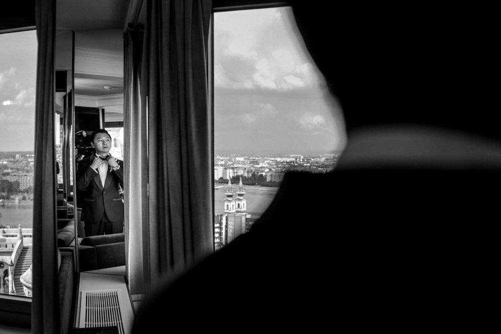 Groom adjusting his tie by a window with a city skyline view in a black and white wedding photo.