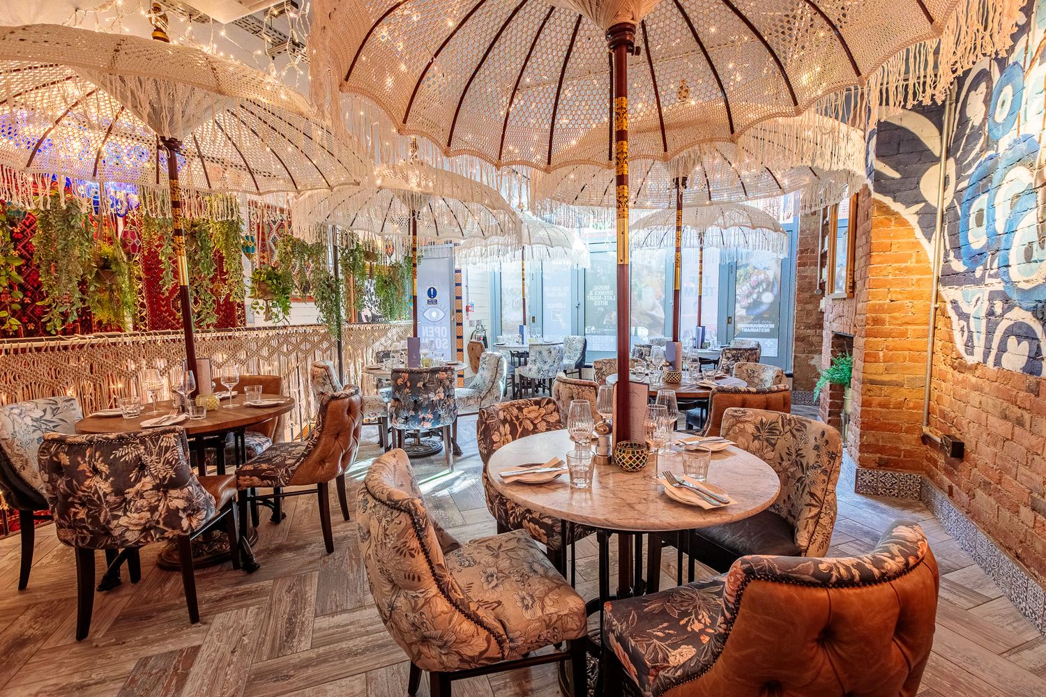 Elegant restaurant interior with ornate white parasols, velvet chairs, brick walls, and lush greenery decor.