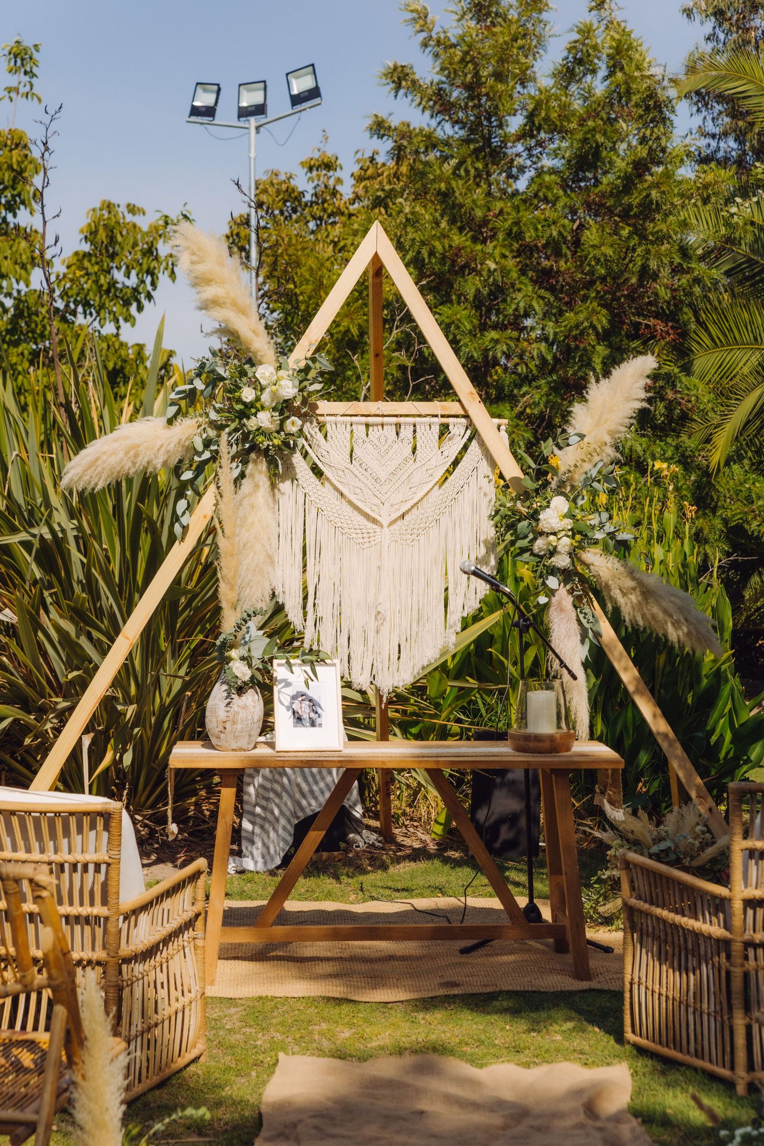 Boho wedding ceremony arch with macrame wall hanging, pampas grass, white flowers, and rustic wooden table.