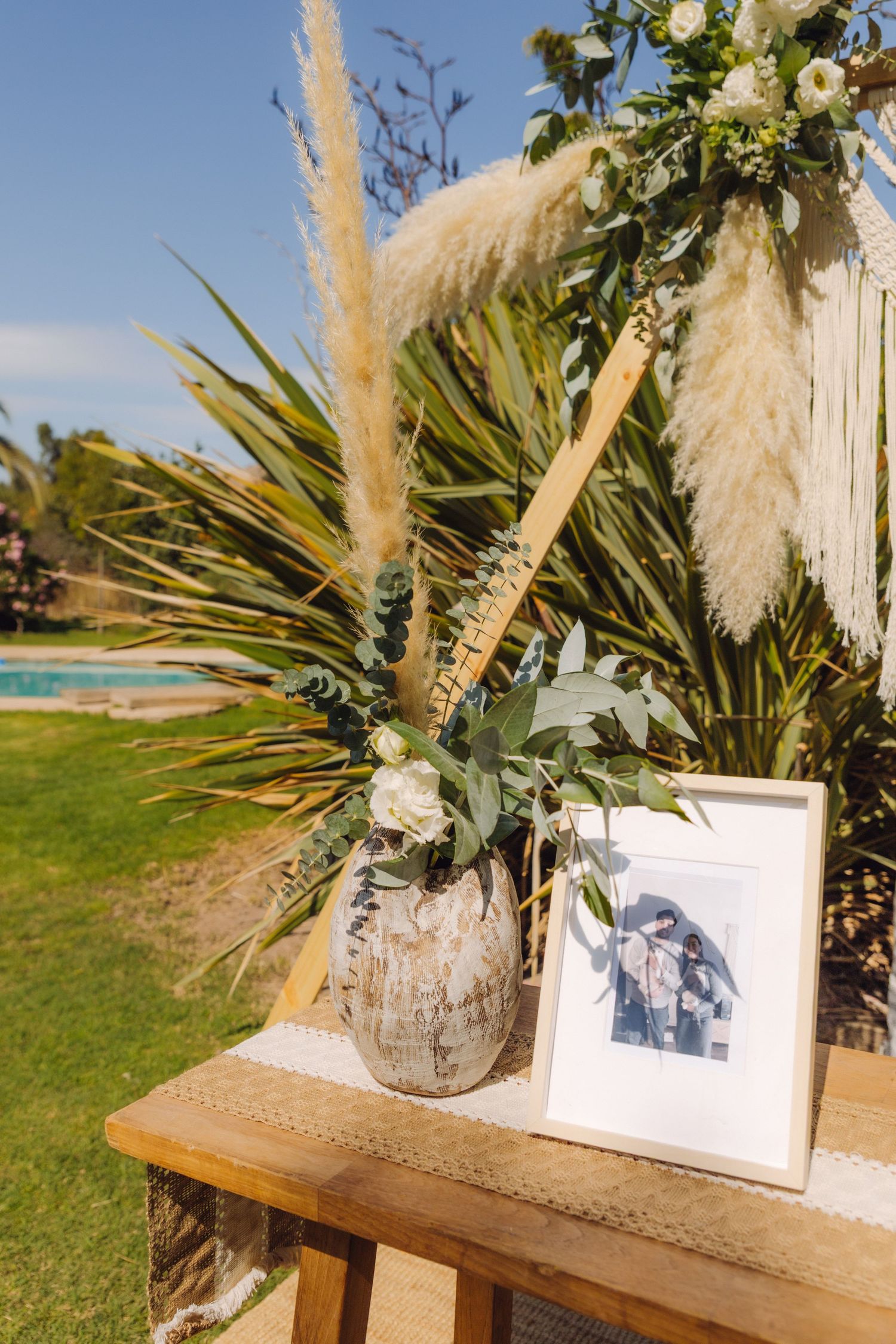 Rustic wooden table with ceramic vase, pampas grass, white roses, and framed photo at boho wedding ceremony.