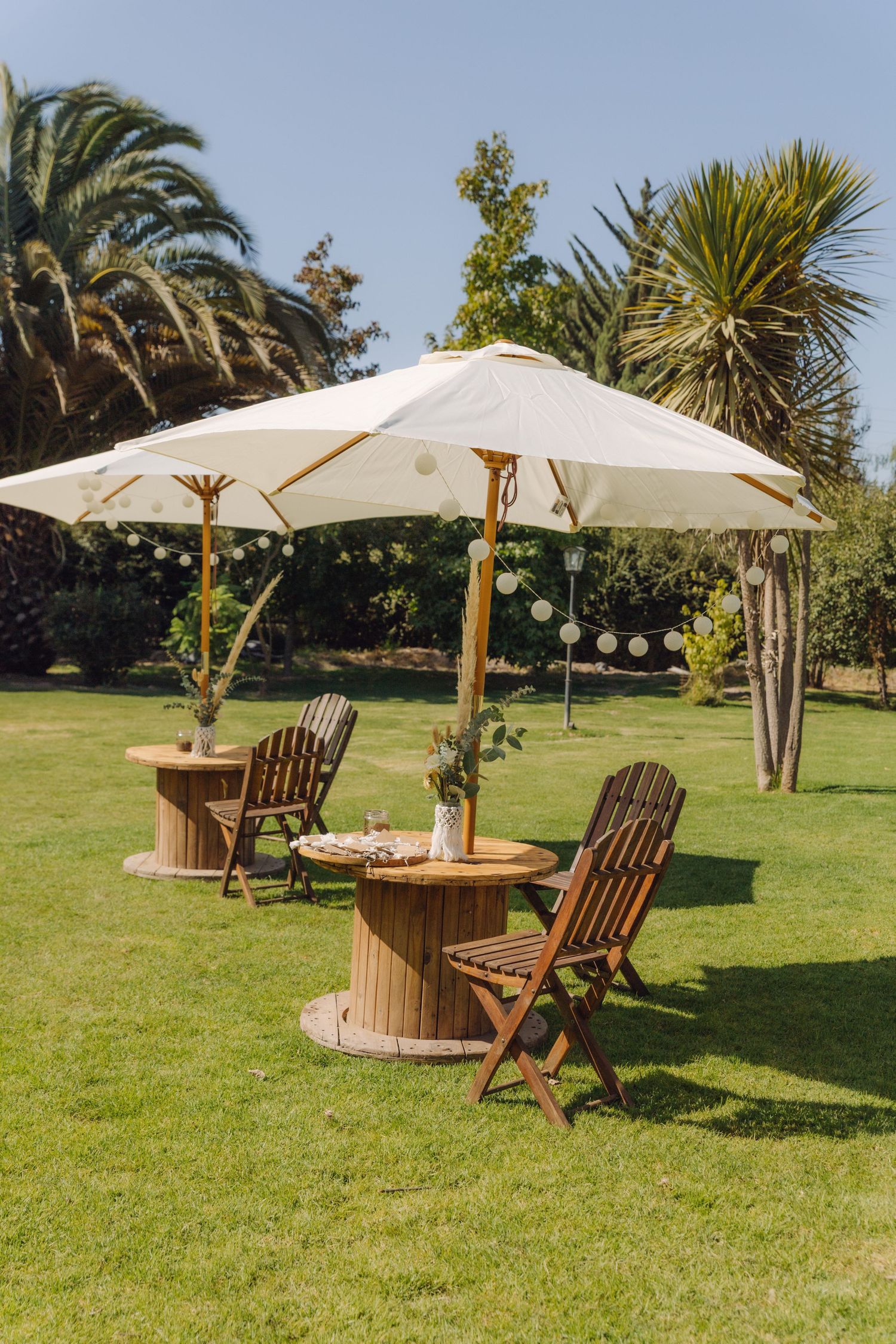 Outdoor garden wedding reception area with wooden spool tables, chairs, white umbrellas, and string lights.